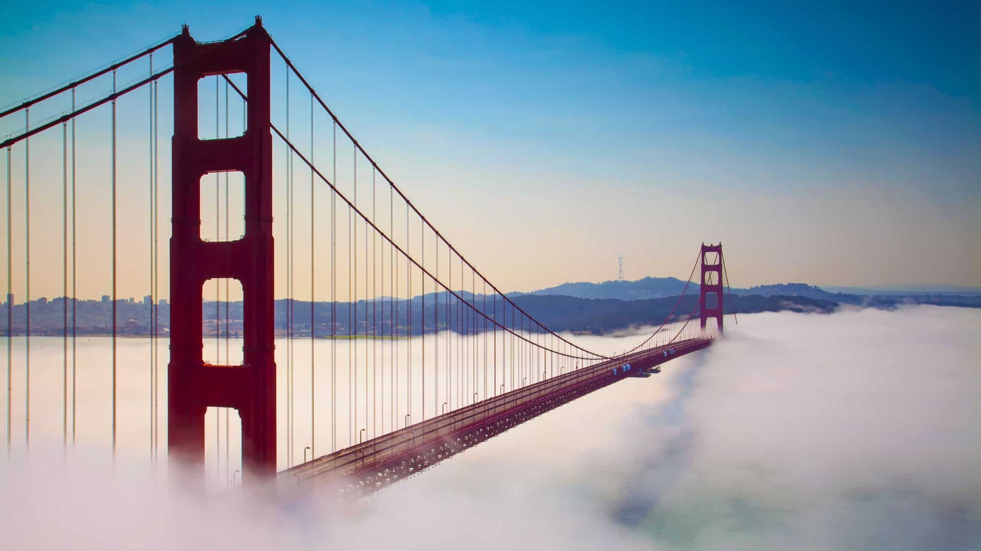 The iconic Golden Gate Bridge in San Francisco rises above a thick layer of coastal fog, with its red towers standing boldly against a clear sky as the city hides below.