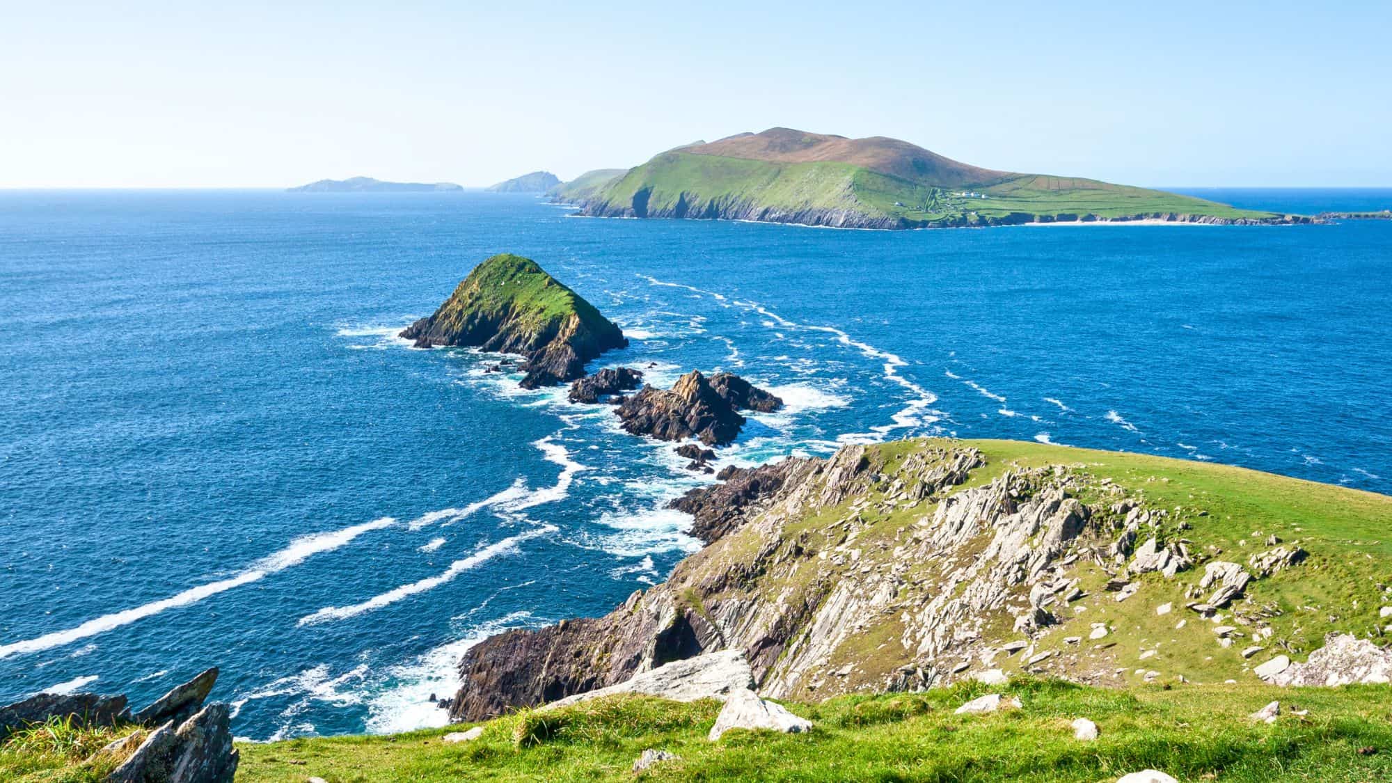 Sweeping ocean view from a grassy cliffside on Great Blasket Island, with jagged rocky outcroppings leading into deep blue Atlantic waters under a clear sky.