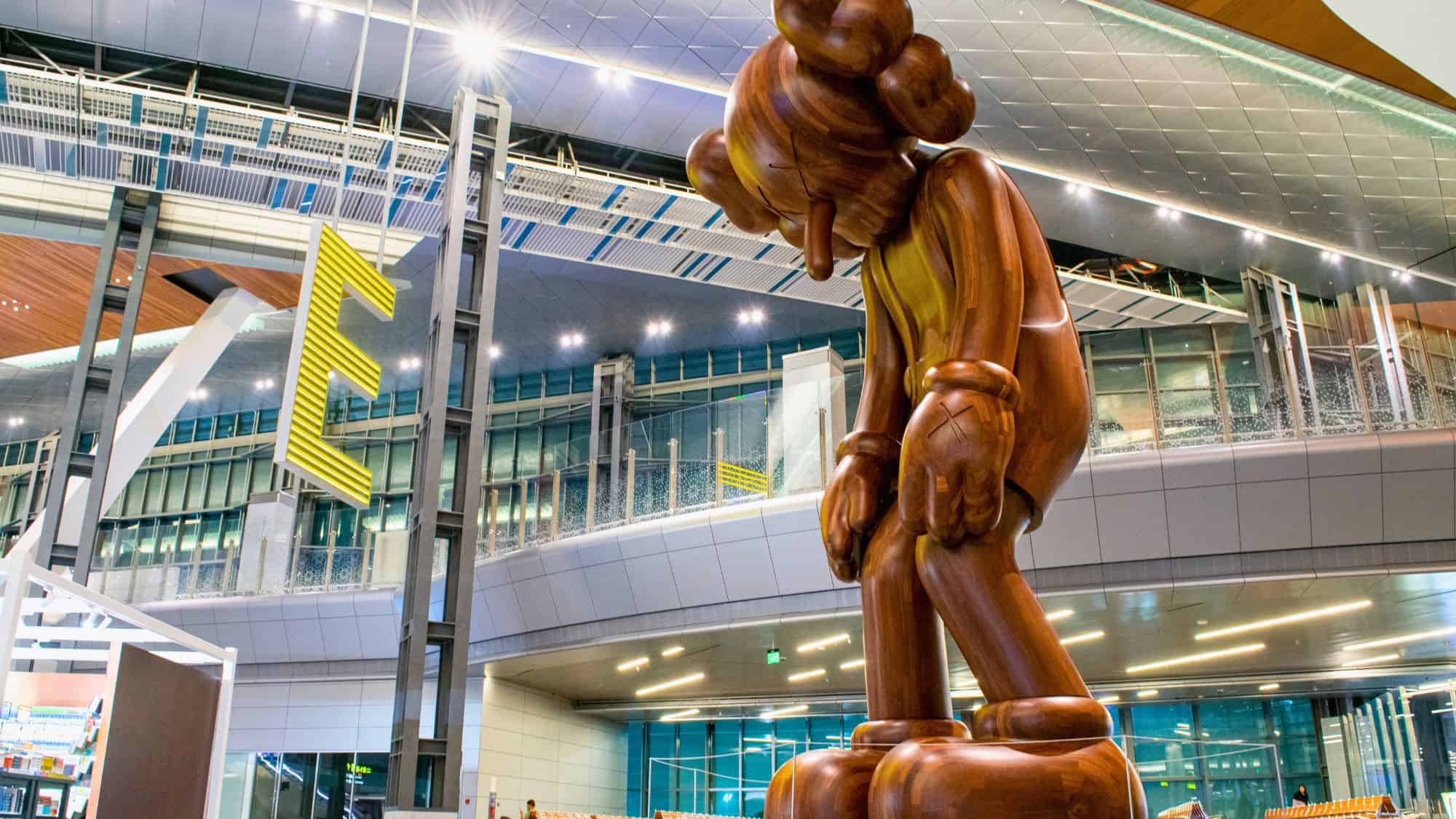 A towering wooden sculpture by artist KAWS stands inside Hamad International Airport, depicting a bowed cartoon-like figure with its hands to its face, illuminated by modern terminal lighting.