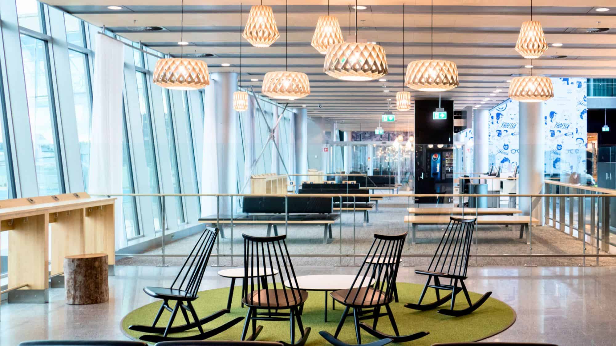 A serene, Scandinavian-style seating area at Helsinki Airport features black rocking chairs, wooden light fixtures, and minimalist furniture framed by large slanted windows and soft natural light.