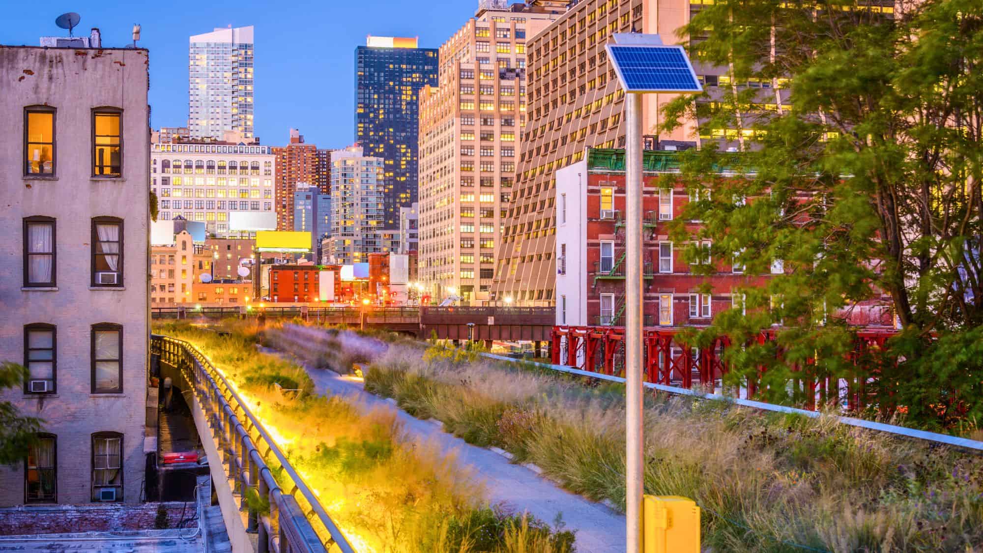 An illuminated view of the High Line in New York City at twilight, with wild grasses lining the elevated path and a backdrop of skyscrapers glowing in the evening light.