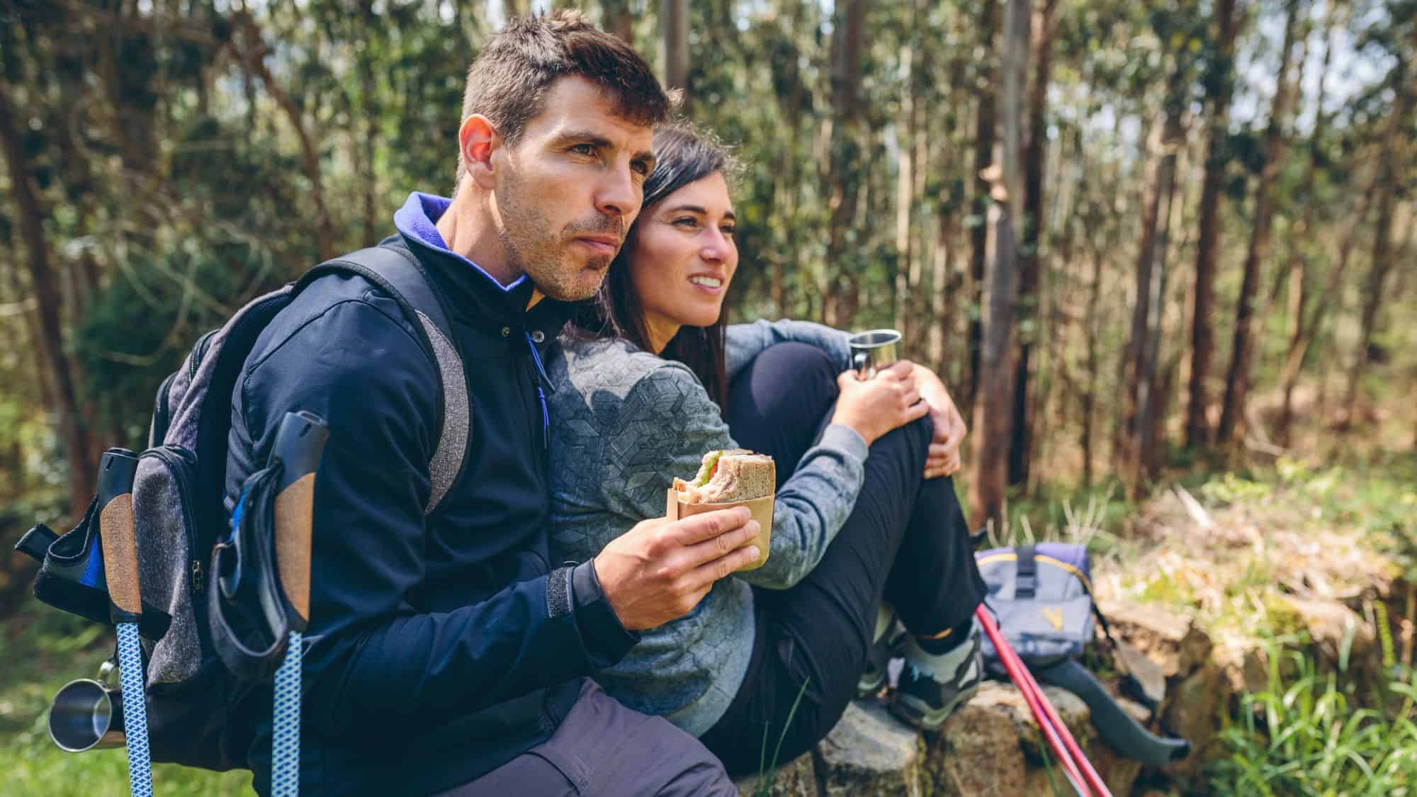 A man and woman rest on a log during a hike, snacking on sandwiches and sipping from metal mugs with trekking poles and a backpack beside them.