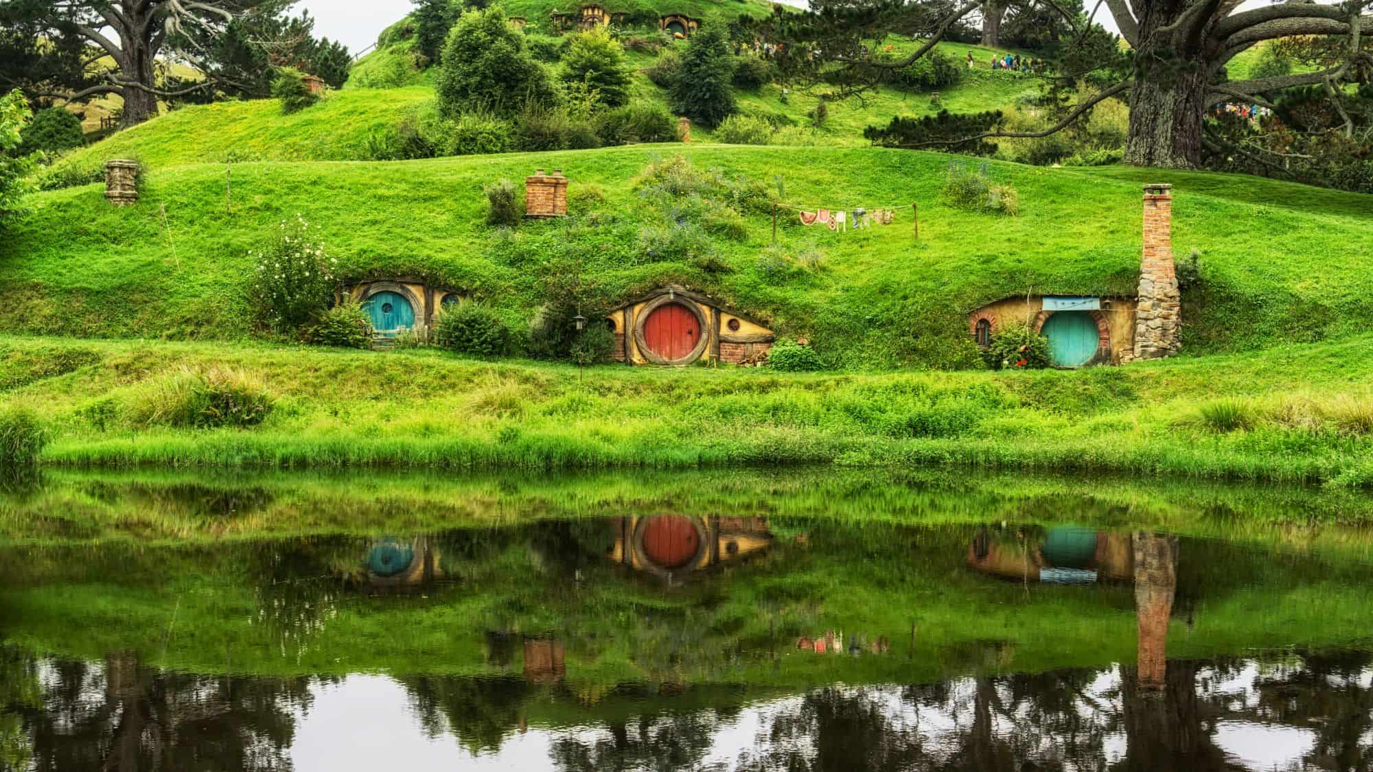 Charming hobbit holes with round doors built into lush green hills, reflected in a still pond, surrounded by flowering bushes and a peaceful, storybook landscape.