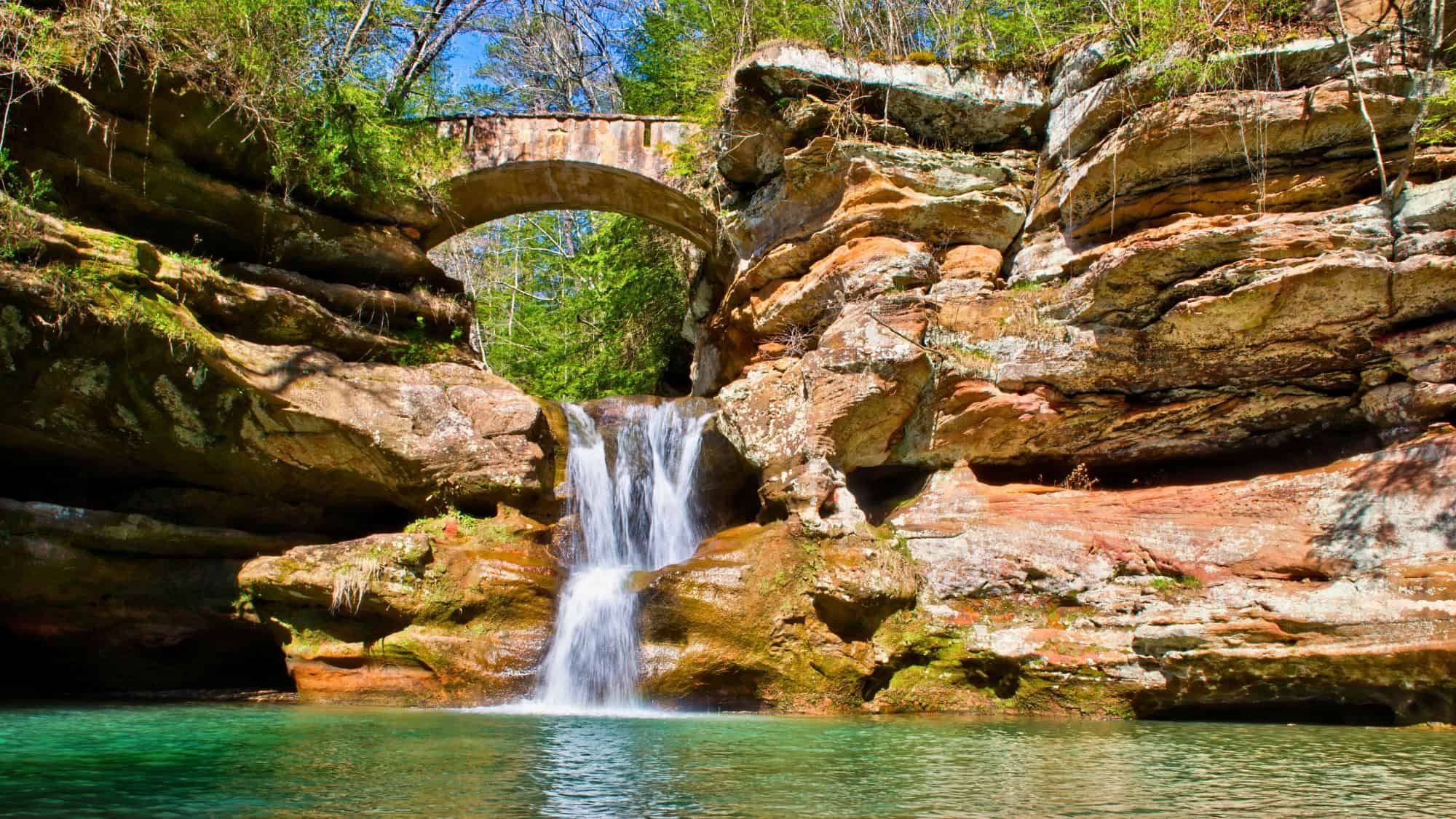 A small waterfall spills beneath a stone arch bridge nestled between red rock cliffs, with a bright turquoise pool below in Hocking Hills State Park.