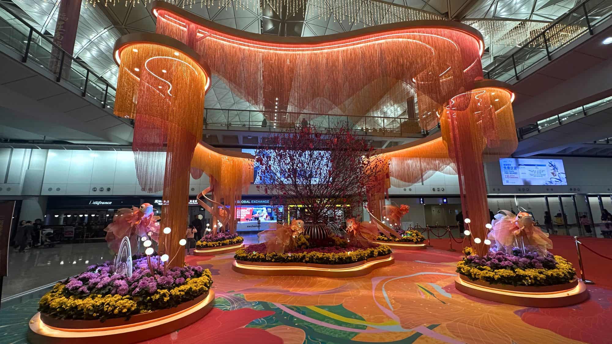 Lavish red and orange decorations for Lunar New Year fill the terminal at Hong Kong International Airport, with cherry blossoms, golden fish, and cascading lights creating a festive floral installation.