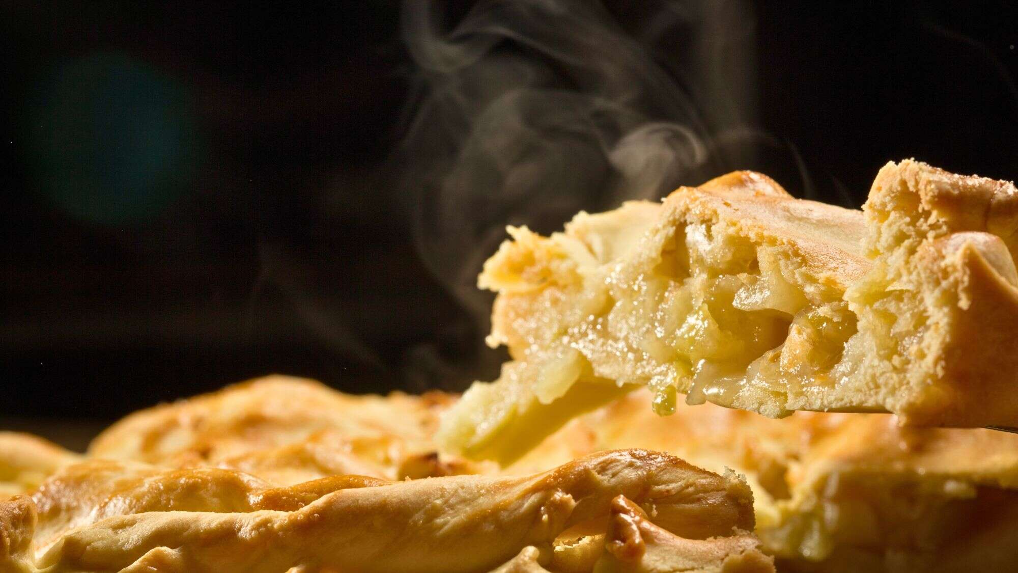 A steaming slice of hot apple pie is lifted from the main pie, showing the gooey apple filling and flaky, golden-brown crust against a dark backdrop.