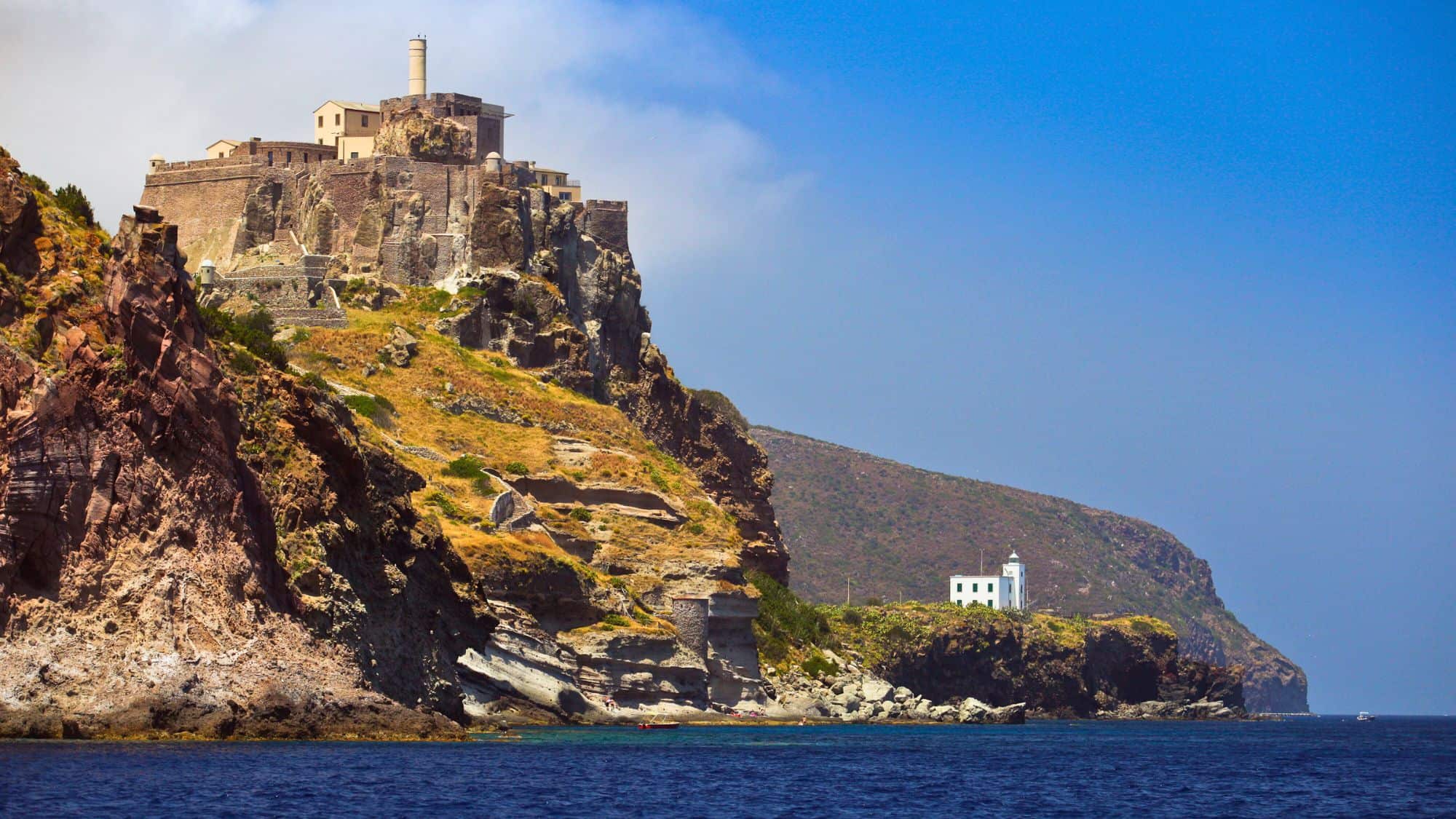 A weathered fortress sits atop steep, rocky cliffs on Isola di Capraia, Italy, with a lighthouse perched at the lower edge, overlooking deep blue Mediterranean waters.