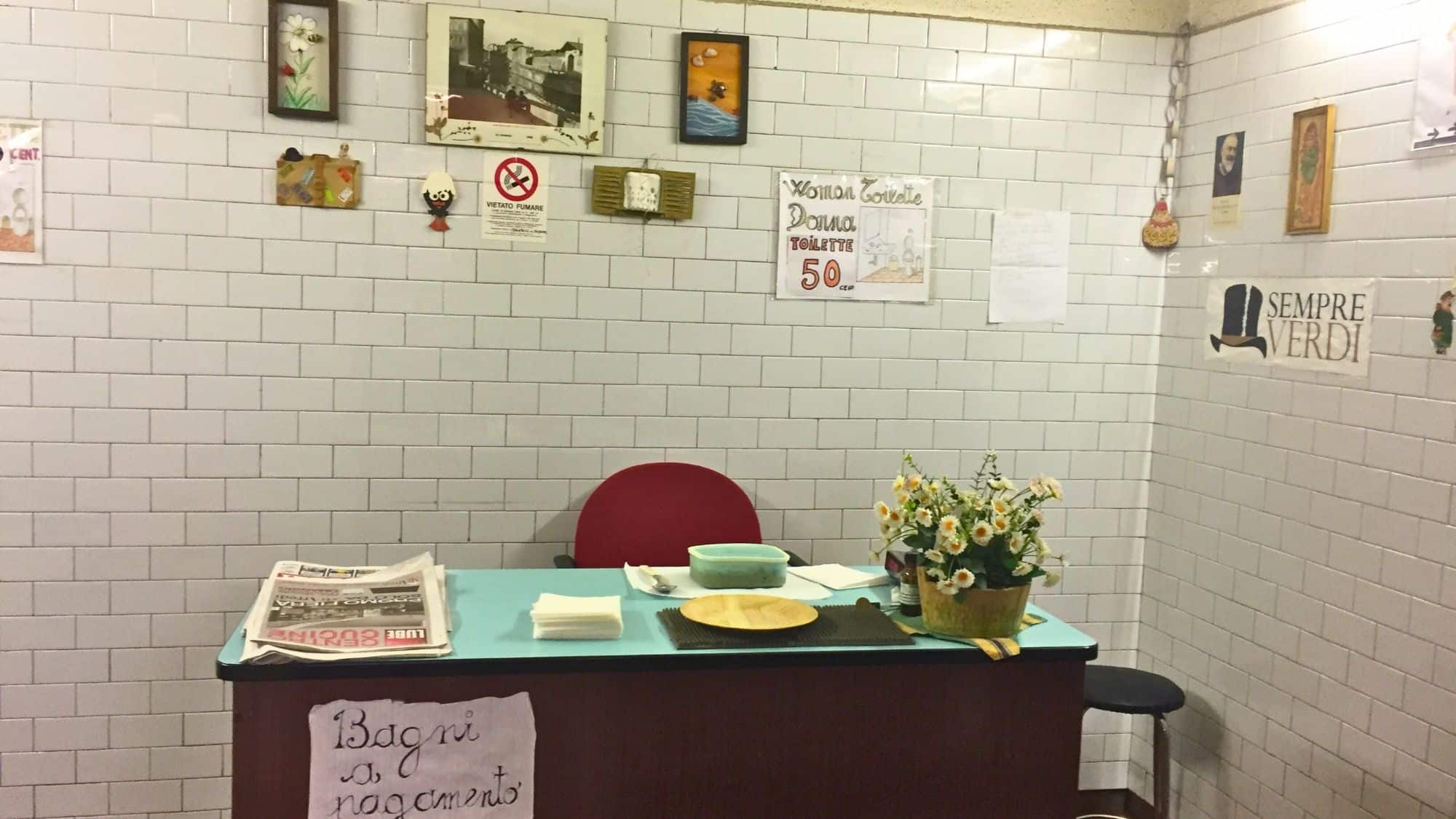 A tiled restroom corner in Italy with a small desk labeled “Bagni a pagamento,” decorated with faux flowers, napkins, a red chair, and various wall signs indicating a paid toilet area.