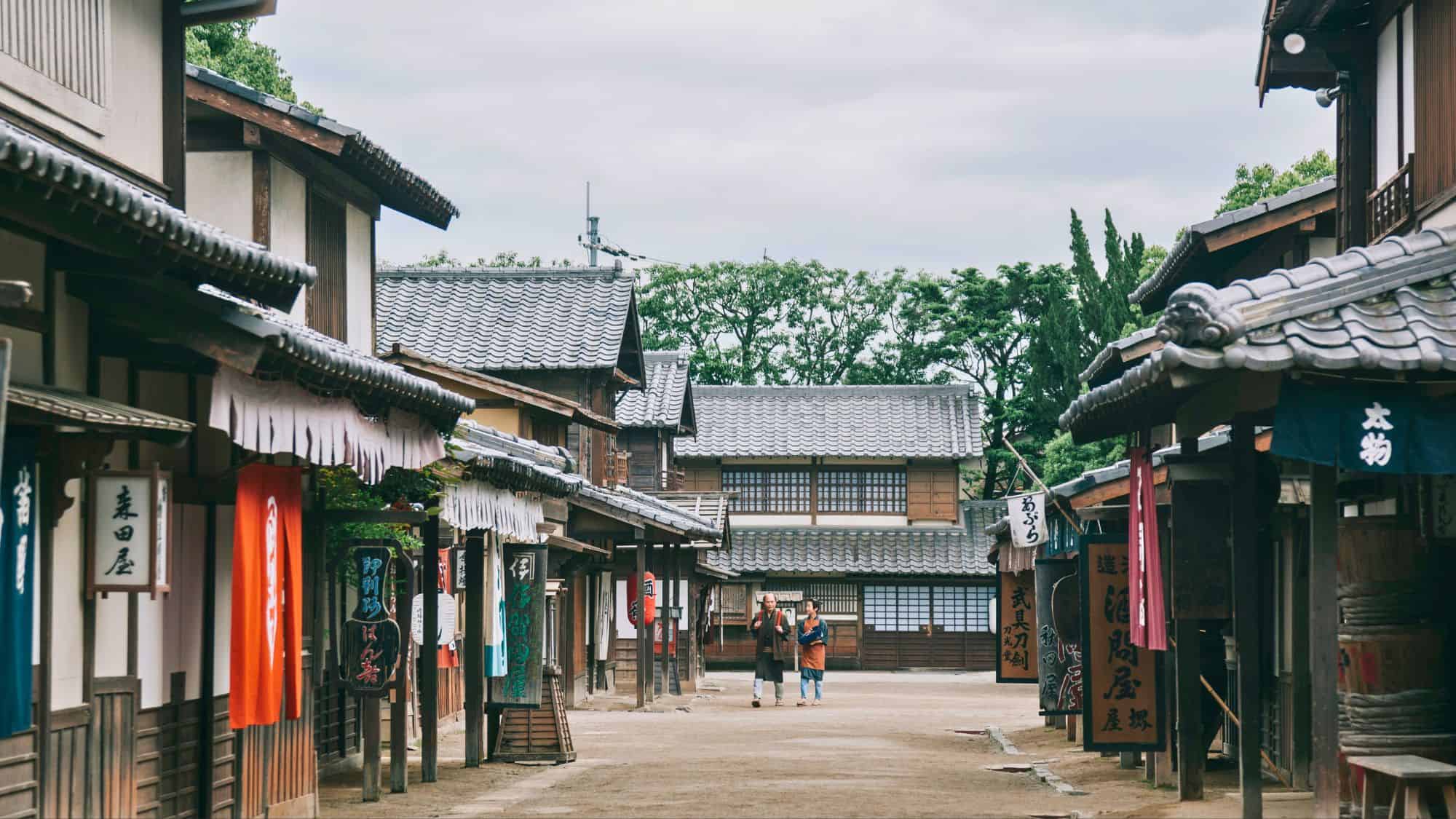 Quiet traditional Japanese street scene with tiled roof buildings and hanging signs, showing two people walking down a dirt path lined with Edo-period architecture.