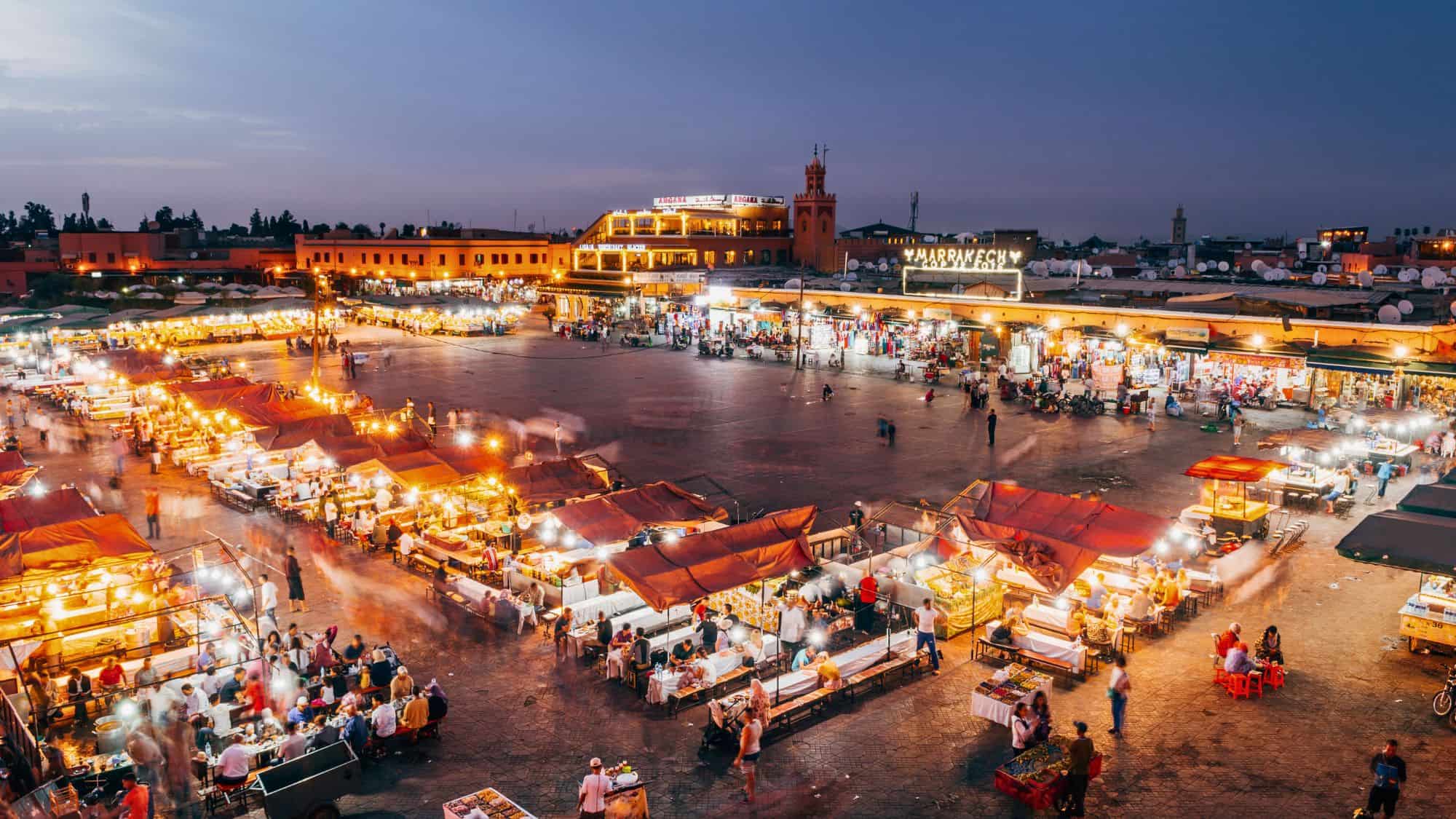 An atmospheric view of Jemaa el-Fnaa square at dusk in Marrakech, Morocco, glowing with warm lights from open-air food stalls as people gather around to eat and shop in the expansive plaza.