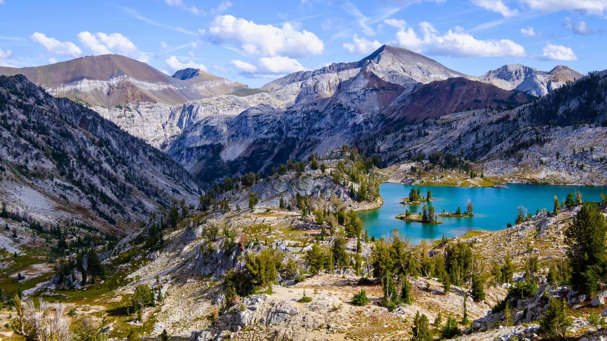 A vibrant alpine landscape near Joseph, Oregon, with a turquoise glacial lake surrounded by rocky mountains and scattered evergreen trees beneath a blue sky with clouds.