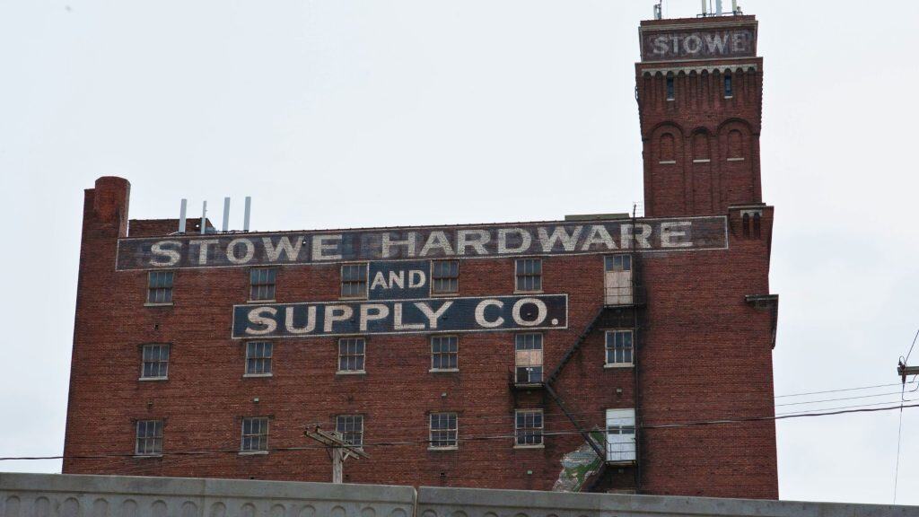 An old red-brick industrial building labeled “Stowe Hardware and Supply Co.” with faded white painted lettering and boarded-up windows, viewed from across a barrier.