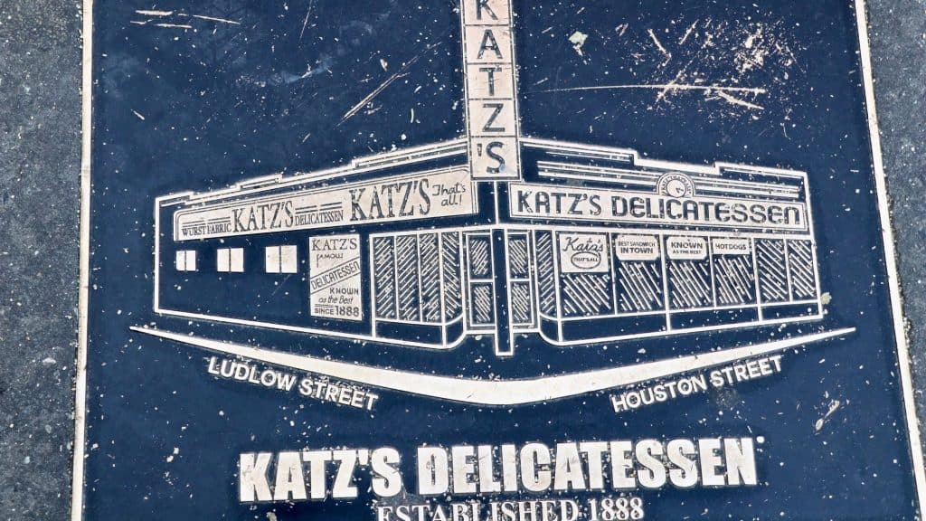 A worn metal plaque embedded in the sidewalk shows an etched drawing of Katz's Delicatessen on the corner of Ludlow and Houston Street in New York City, celebrating its establishment in 1888.