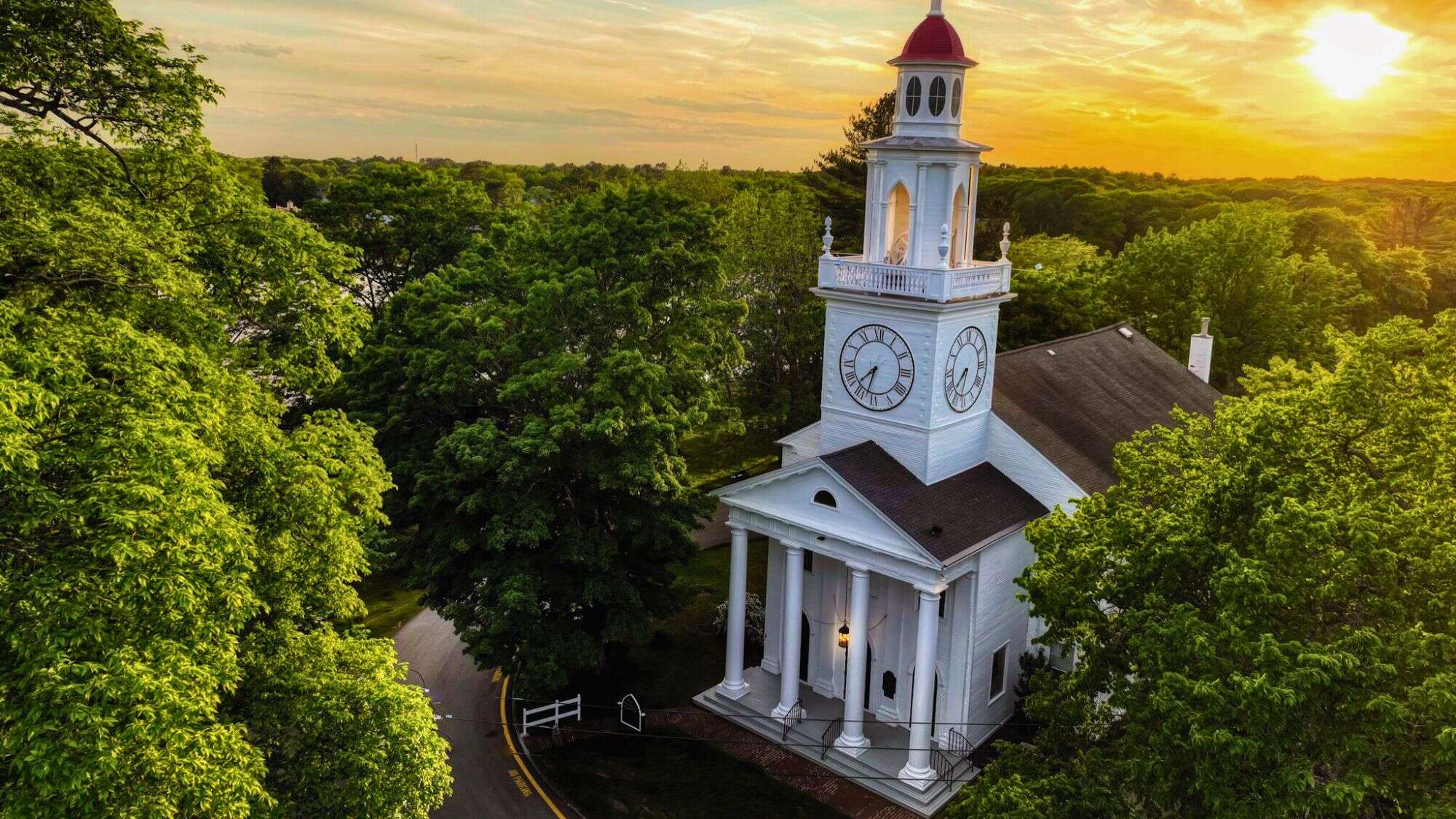 A white colonial-style church with a red cupola and clock tower rises above lush green trees at sunset in Kennebunkport, Maine, glowing with warm golden light.
