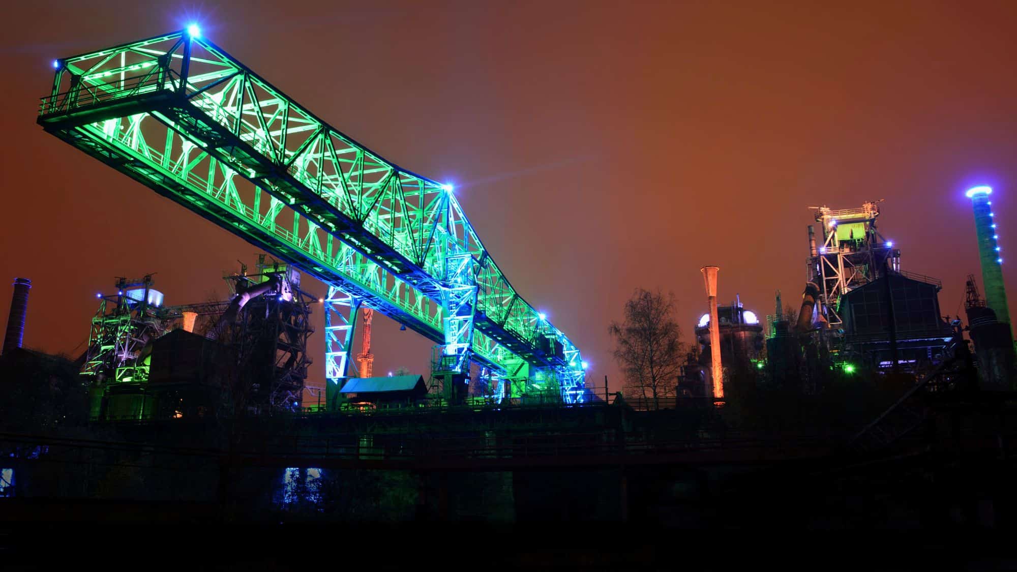 A former industrial plant in Germany, lit up in vivid blue and green lights against an orange sky, featuring towering steel structures and a long elevated bridge.