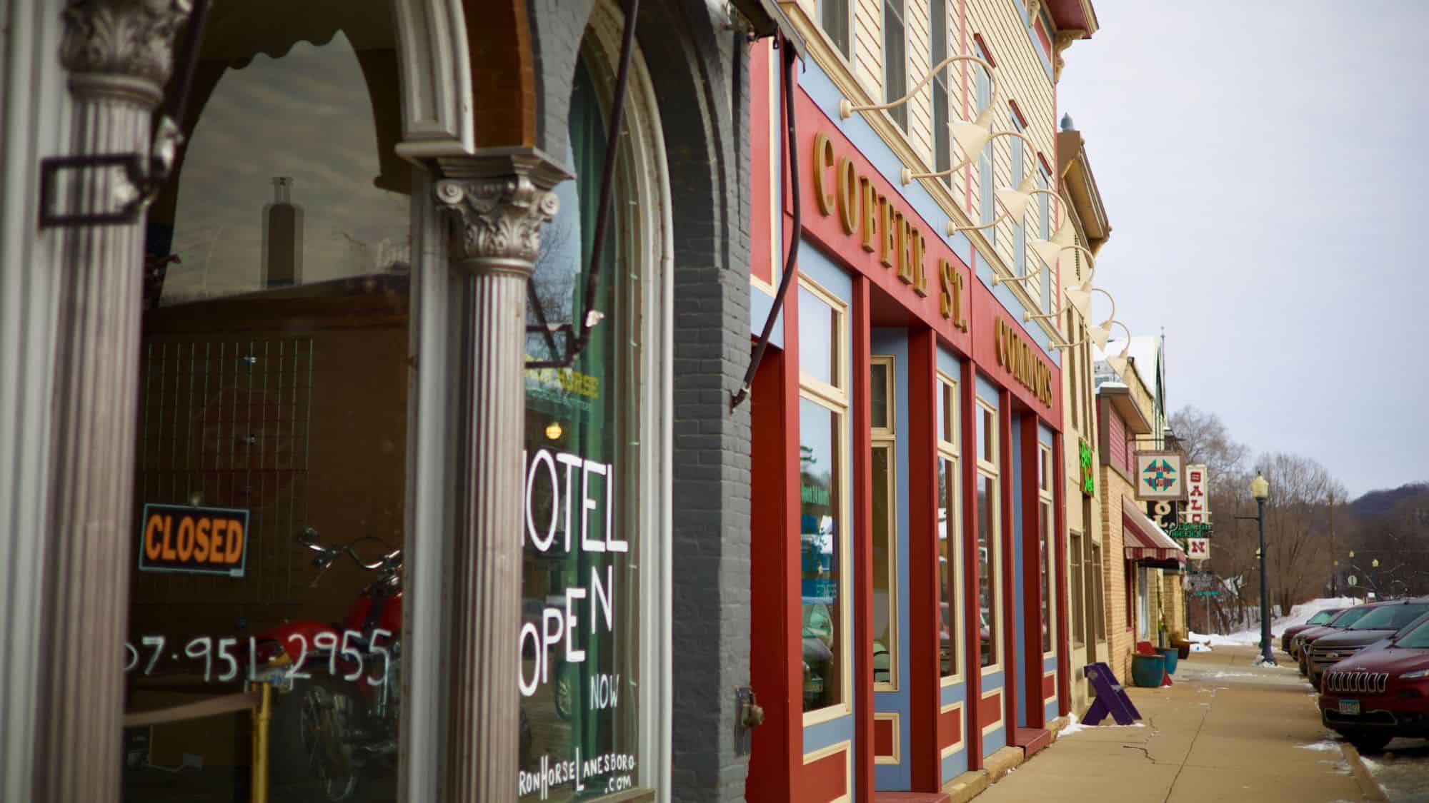 A colorful street scene in downtown Lanesboro, Minnesota, with charming storefronts including Coffee St. Commons, a hotel window sign, and snow-lined sidewalks on a quiet winter day.