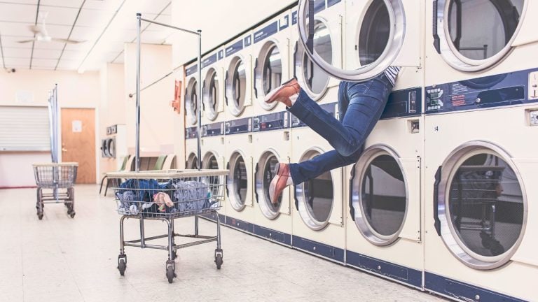 A humorous scene in a laundromat where a person is halfway inside a front-loading dryer, legs sticking out while wearing jeans and brown shoes, next to a laundry cart filled with clothes.