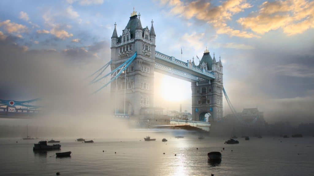 London’s Tower Bridge emerges through dense fog at dawn, lit dramatically from behind as moored boats rest on the River Thames, adding to the serene mystery.