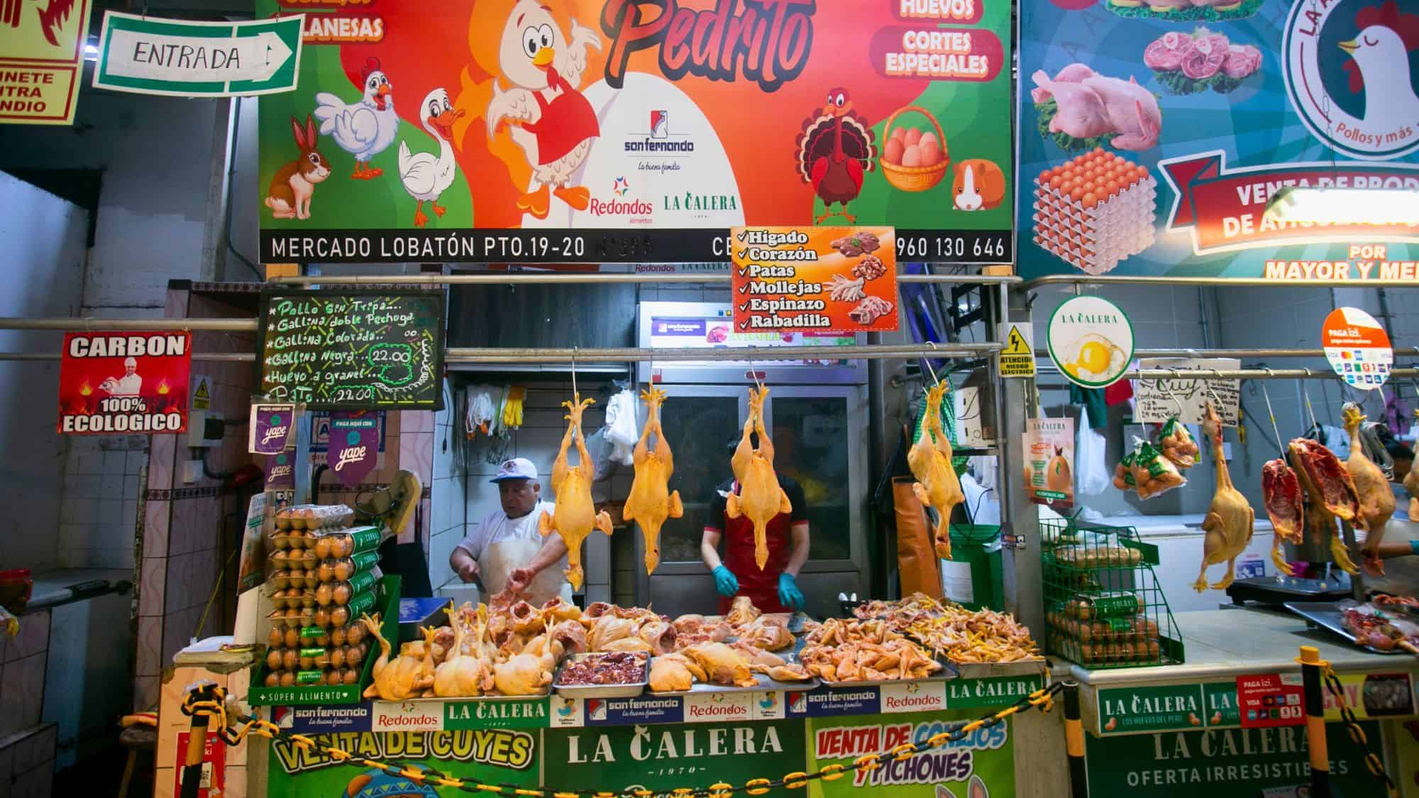 A Peruvian butcher stall at Surquillo Market showcasing whole plucked chickens hanging and cut poultry parts neatly arranged, with colorful signs listing items like liver, gizzards, and wings.