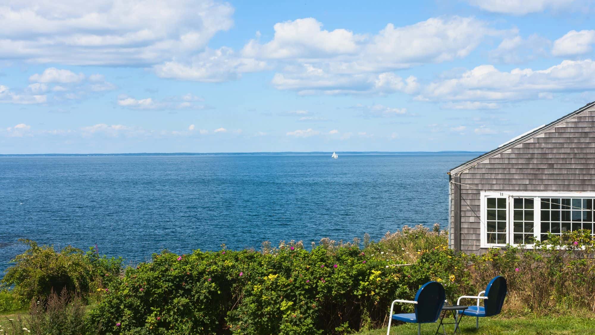 A peaceful coastal scene from Monhegan Island, Maine, showing a shingled seaside cottage, blue lawn chairs, and a wide expanse of calm ocean stretching to the horizon.