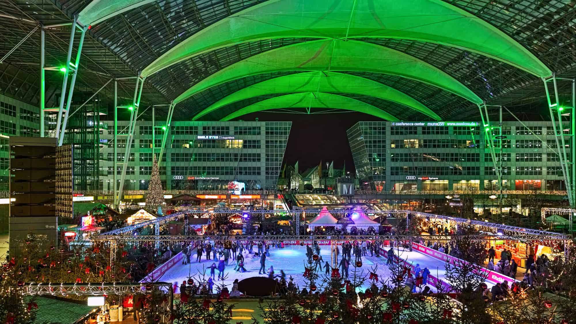 A lively Christmas market with twinkling lights and a large skating rink fills the covered open-air space of Munich Airport, complete with decorated trees and festive stalls under a glowing green roof canopy.