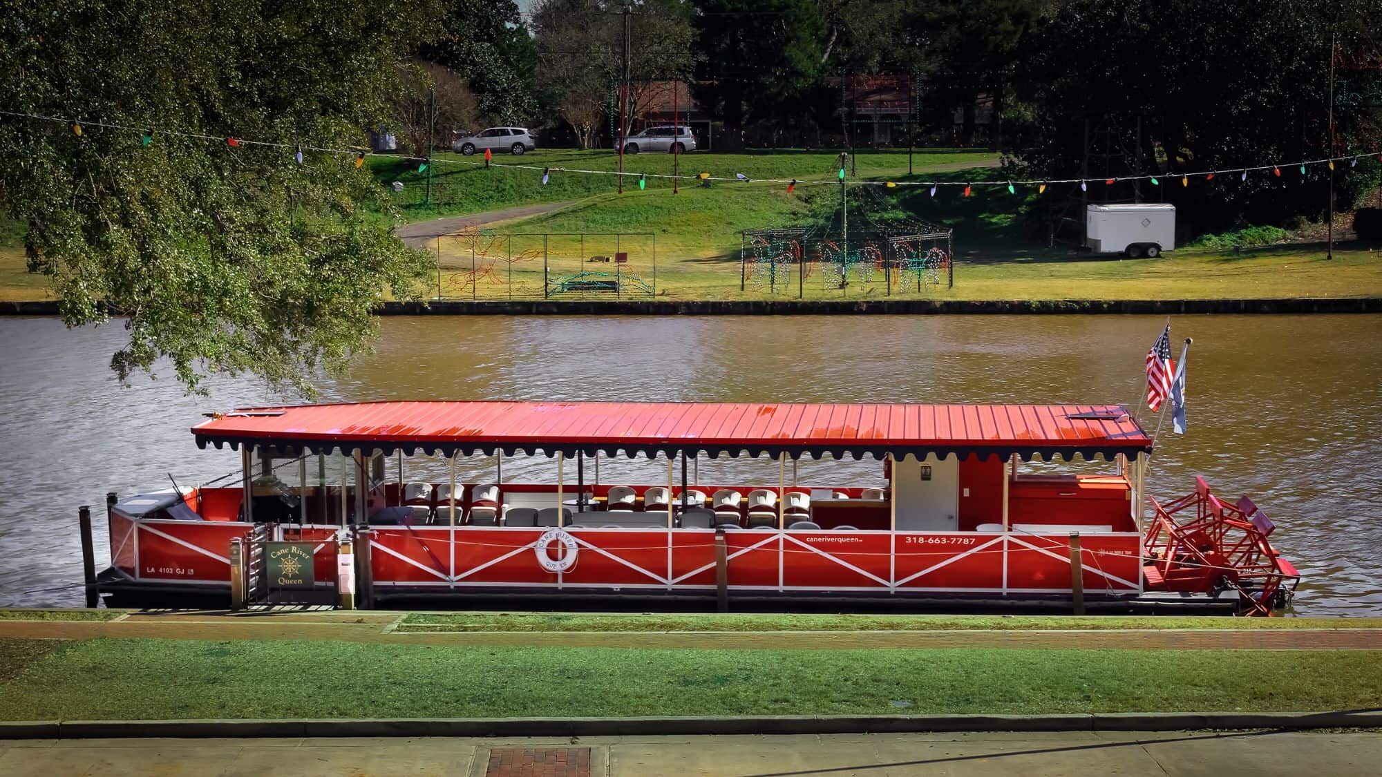 The Cane River Queen, a bright red paddleboat docked on the riverbank in Natchitoches, Louisiana, with string lights overhead and American flags flying on a sunny day.