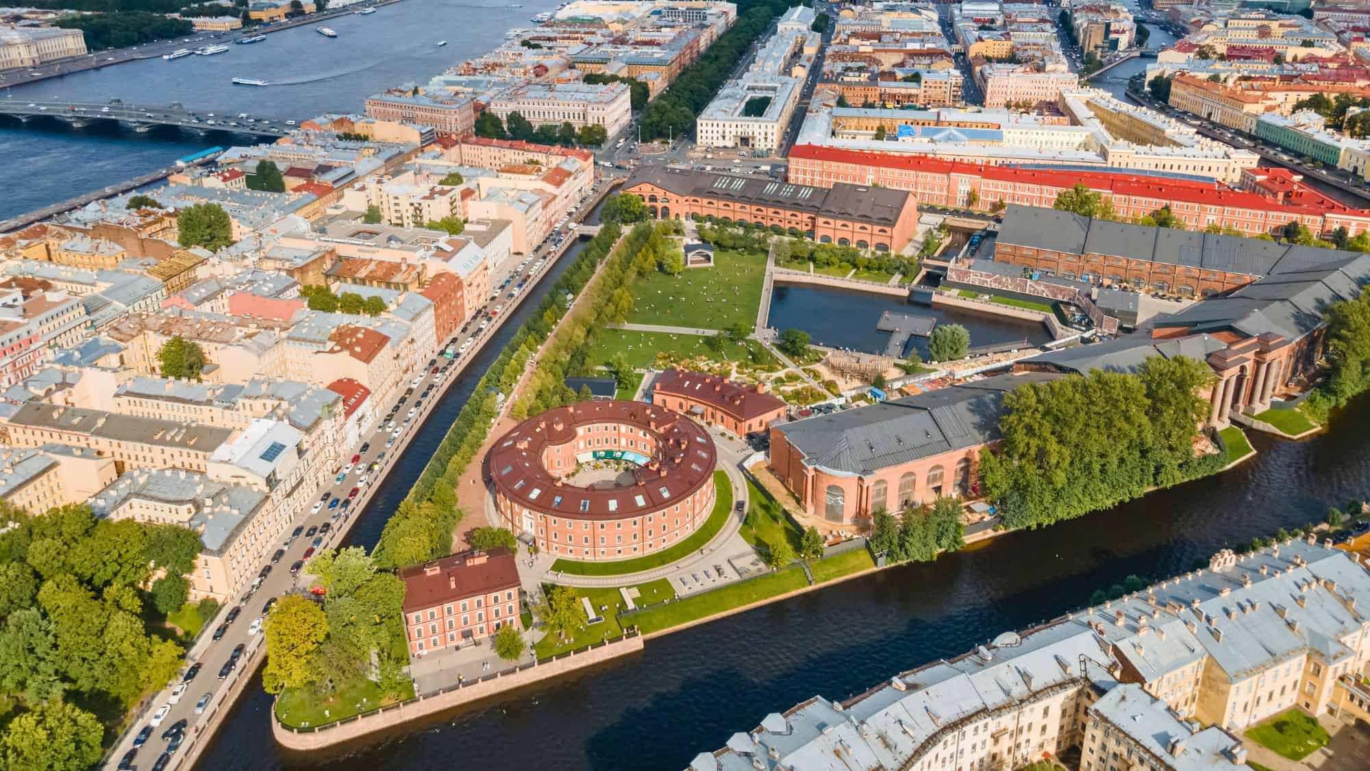 Aerial view of New Holland Island in St. Petersburg, Russia, with a circular red-roofed building, green public park space, and a canal winding through the urban landscape.