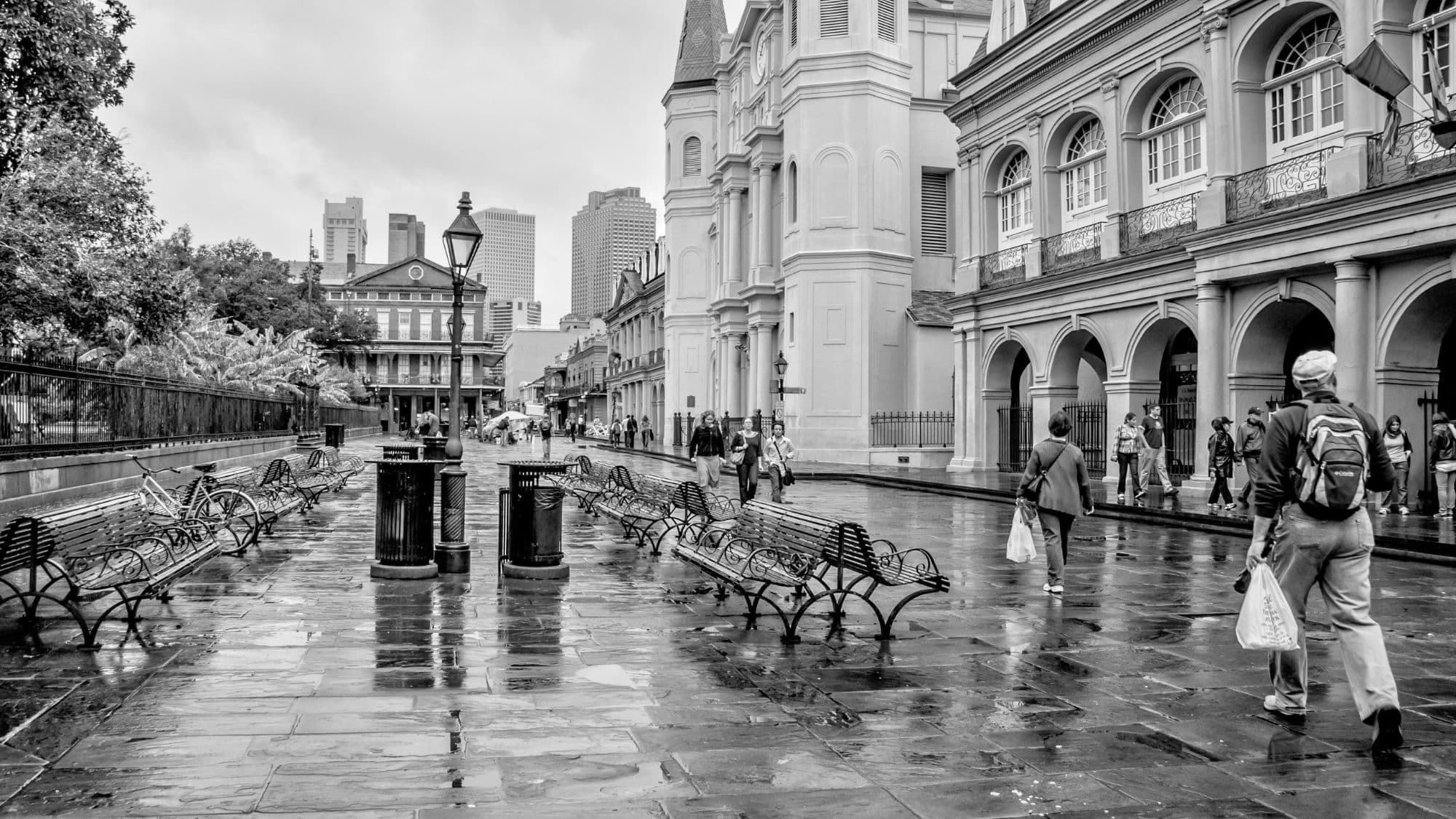 A black-and-white photo of Jackson Square in New Orleans shows wet cobblestones glistening after rain, with pedestrians strolling past the St. Louis Cathedral and classic French Quarter buildings.