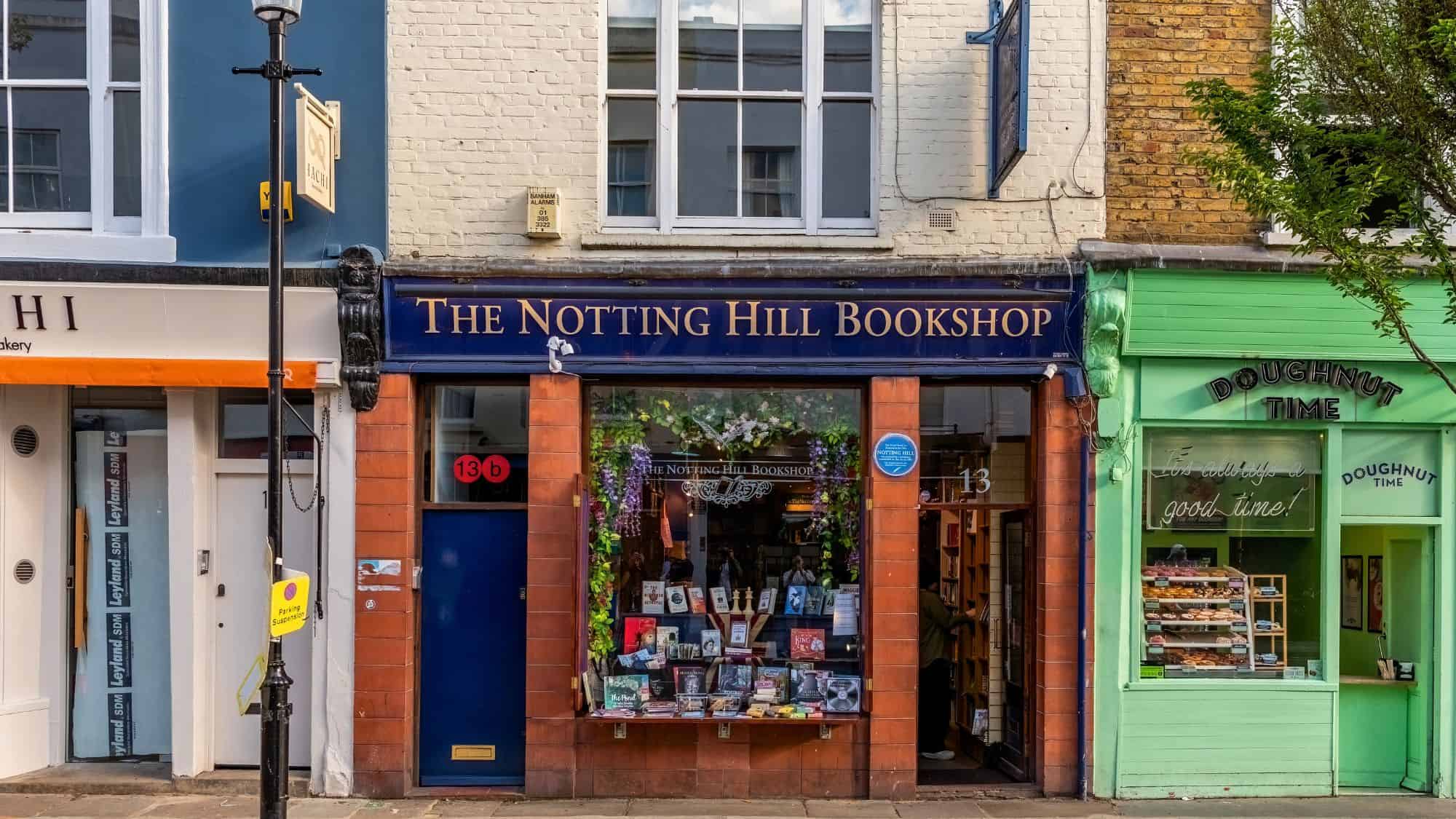 The quaint exterior of The Notting Hill Bookshop in London, with a cozy display window of books framed by red brick and flanked by pastel storefronts on a charming street.