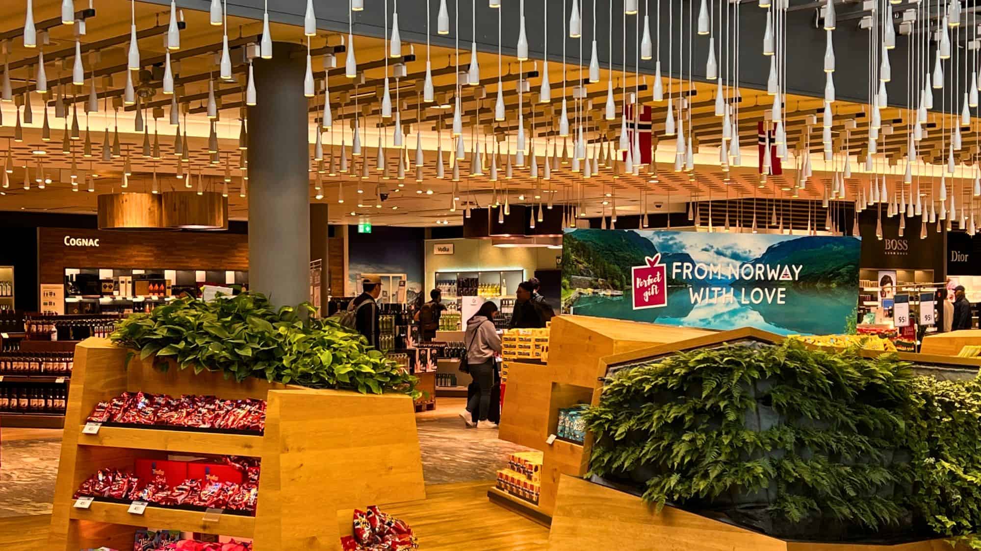 A cozy and modern duty-free section at Oslo Airport featuring wooden displays filled with snacks and greenery, hanging pendant lights, and a sign that reads “From Norway with Love.”