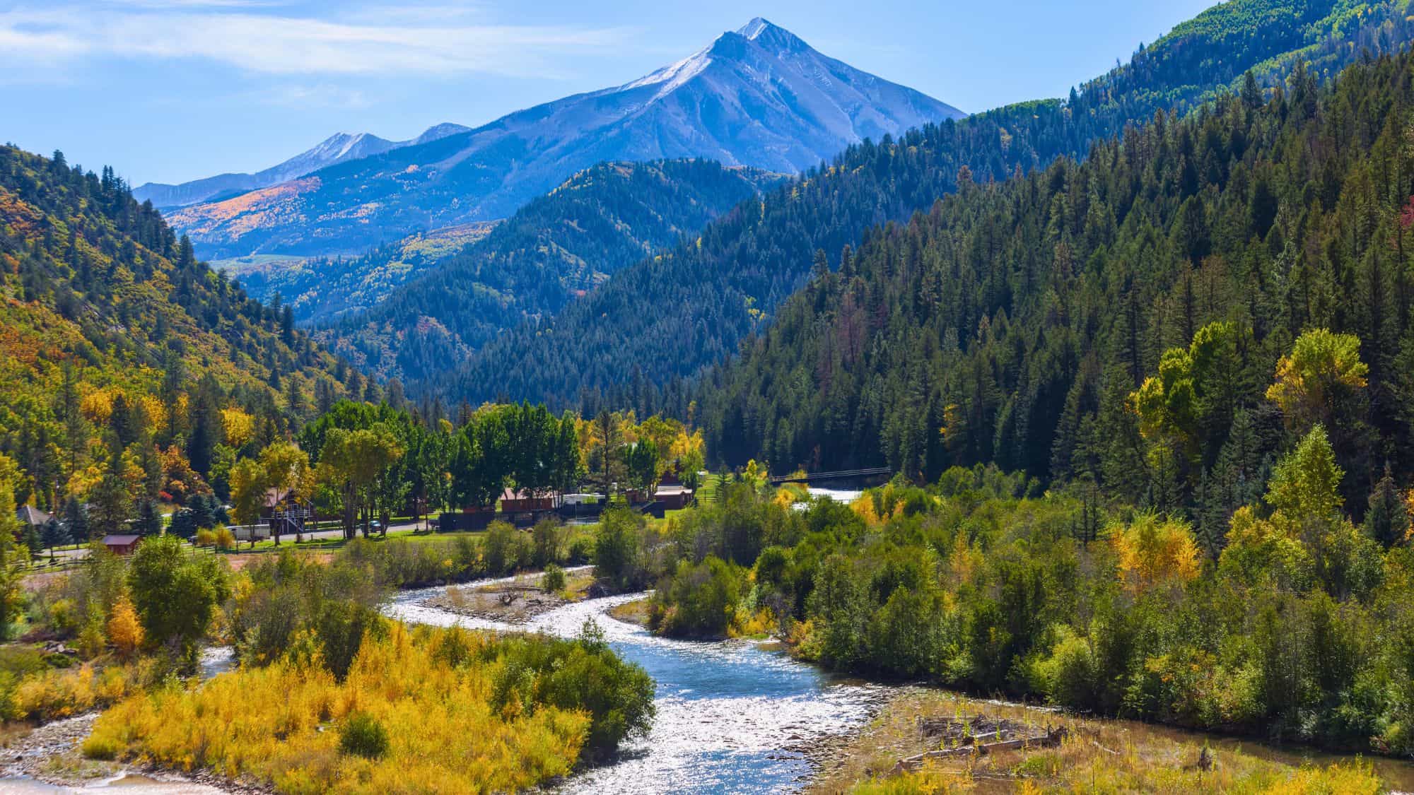 Colorful fall foliage surrounds a flowing river in Paonia, Colorado, with tree-covered hills and a snow-capped mountain rising in the distance under a clear blue sky.