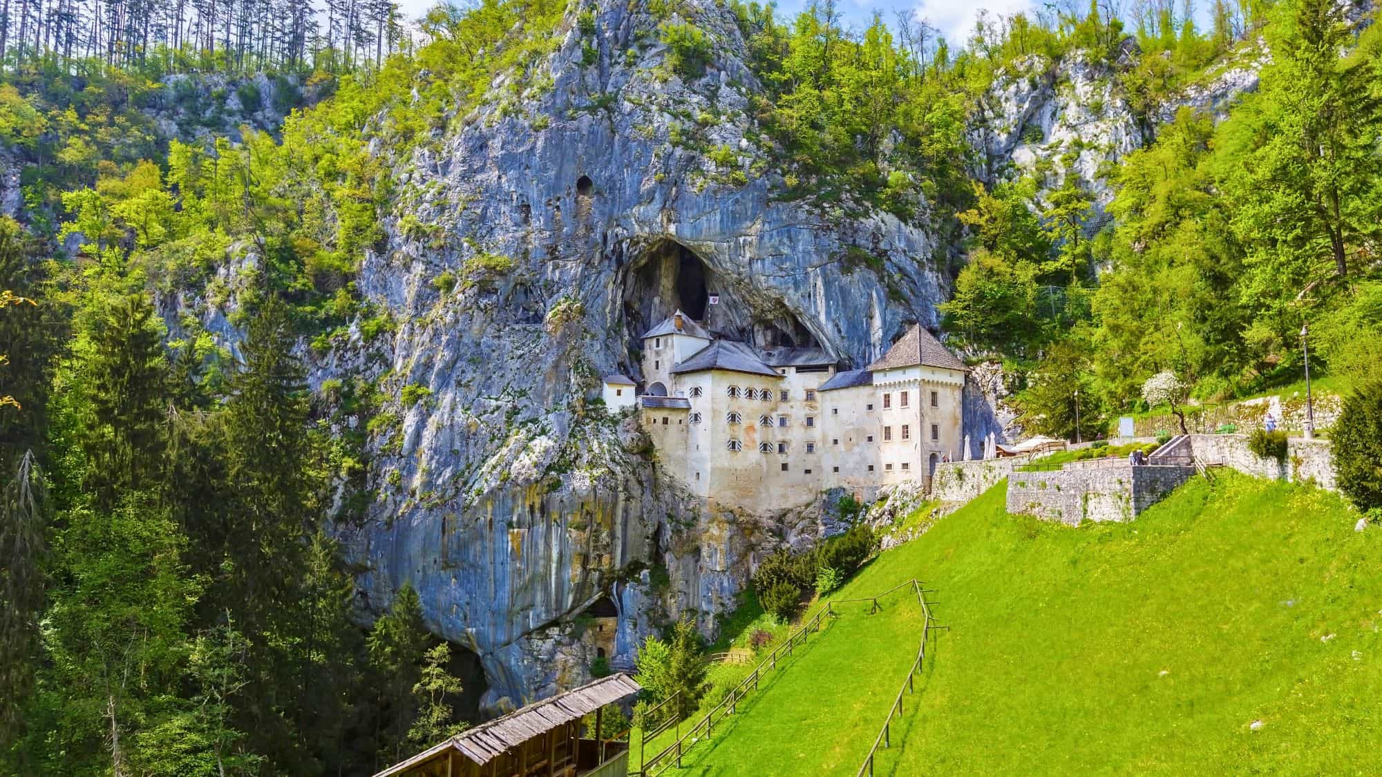 A stone castle built into the mouth of a rocky cave and nestled into a lush, forested hillside in Slovenia. The historic structure blends dramatically with the rugged limestone cliffs, surrounded by green trees and a sloping grassy pasture.