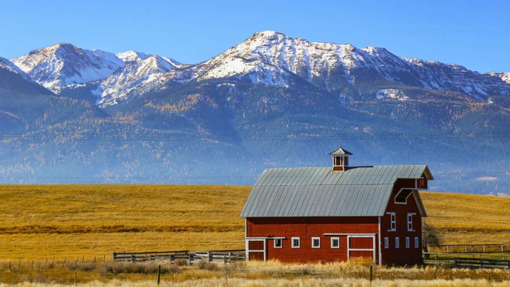 A striking red barn with a silver roof stands in a golden pasture near Joseph, Oregon, with snow-capped peaks of the Wallowa Mountains rising dramatically in the background.