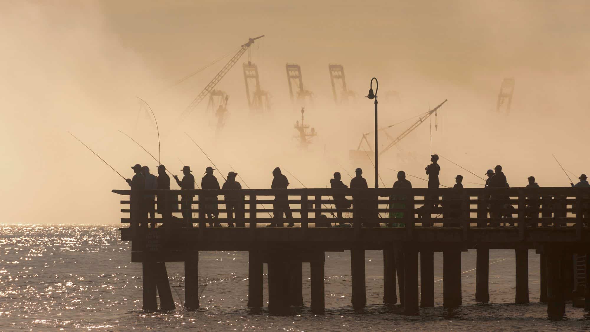 Silhouettes of people line a wooden pier in the early morning mist, casting fishing rods into the shimmering waters of Elliott Bay as towering shipyard cranes loom faintly in the fog behind them.