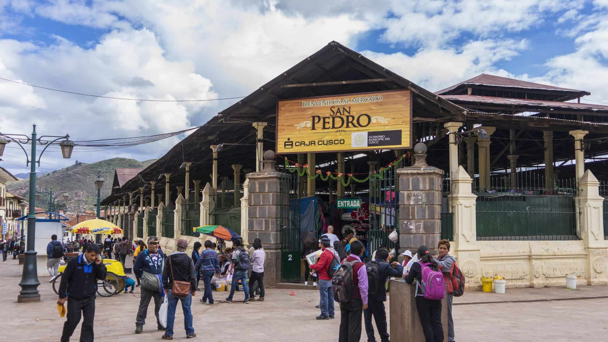 Street view of San Pedro Market in Cusco, Peru, with locals and tourists gathered outside its gated entrance under a wooden sign reading “Bienvenidos al Mercado San Pedro,” set against a backdrop of hills and cloudy skies.