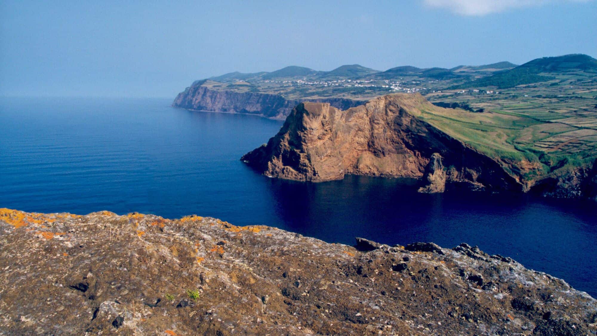 Dramatic cliffs drop into the Atlantic Ocean from the edge of São Jorge island in the Azores, with patchwork fields and distant hills stretching along the ridgeline.
