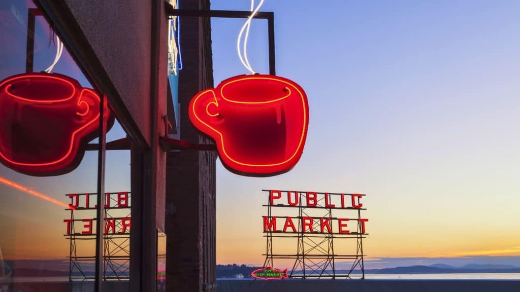 A bright red neon coffee cup sign glows above Seattle’s iconic Public Market at sunset, with its reflection mirrored in a nearby window and the waterfront visible in the background.