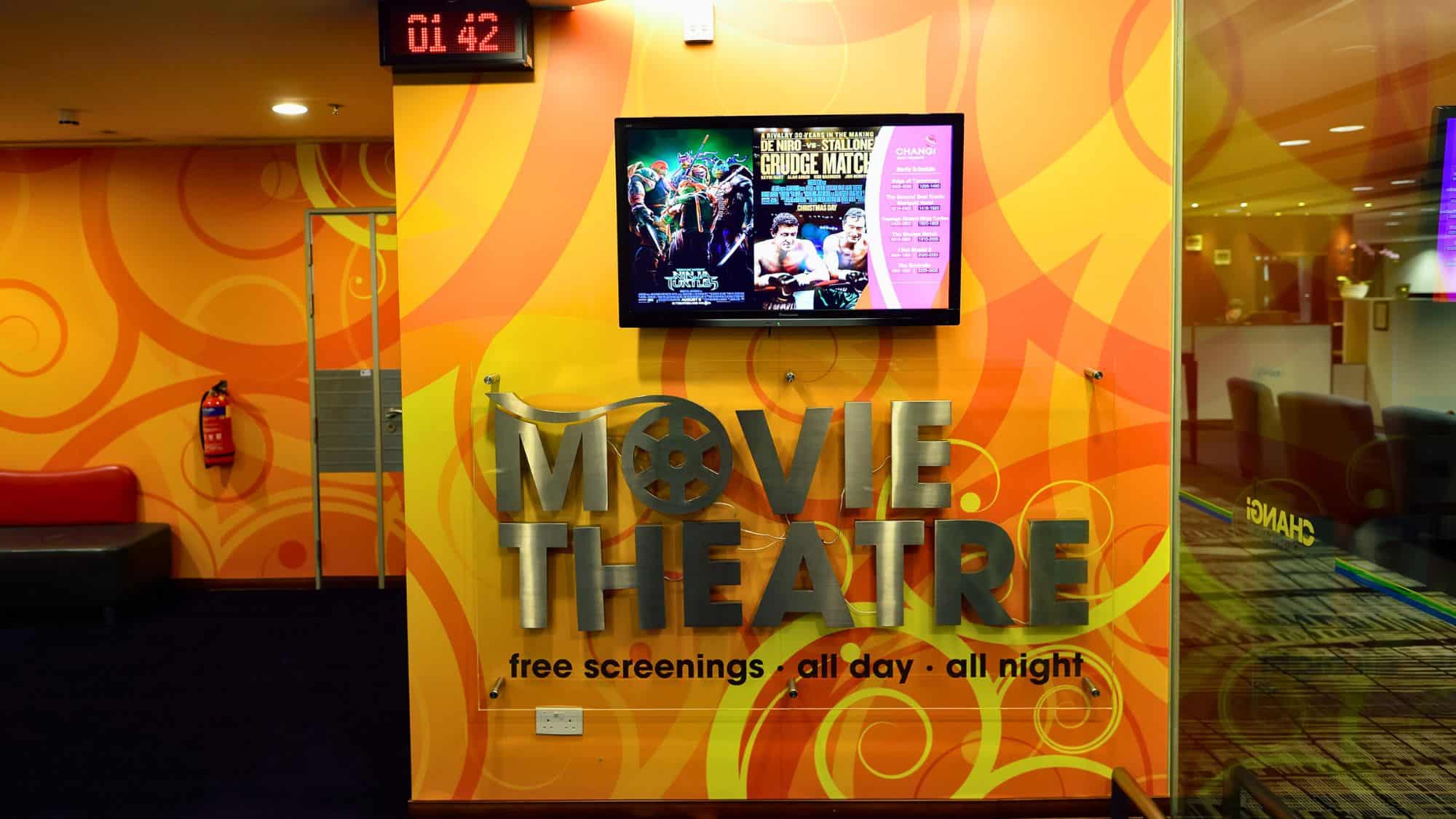 A cozy, brightly decorated airport movie theatre with a sign offering free screenings all day and night, featuring posters and an LED clock above the entrance.
