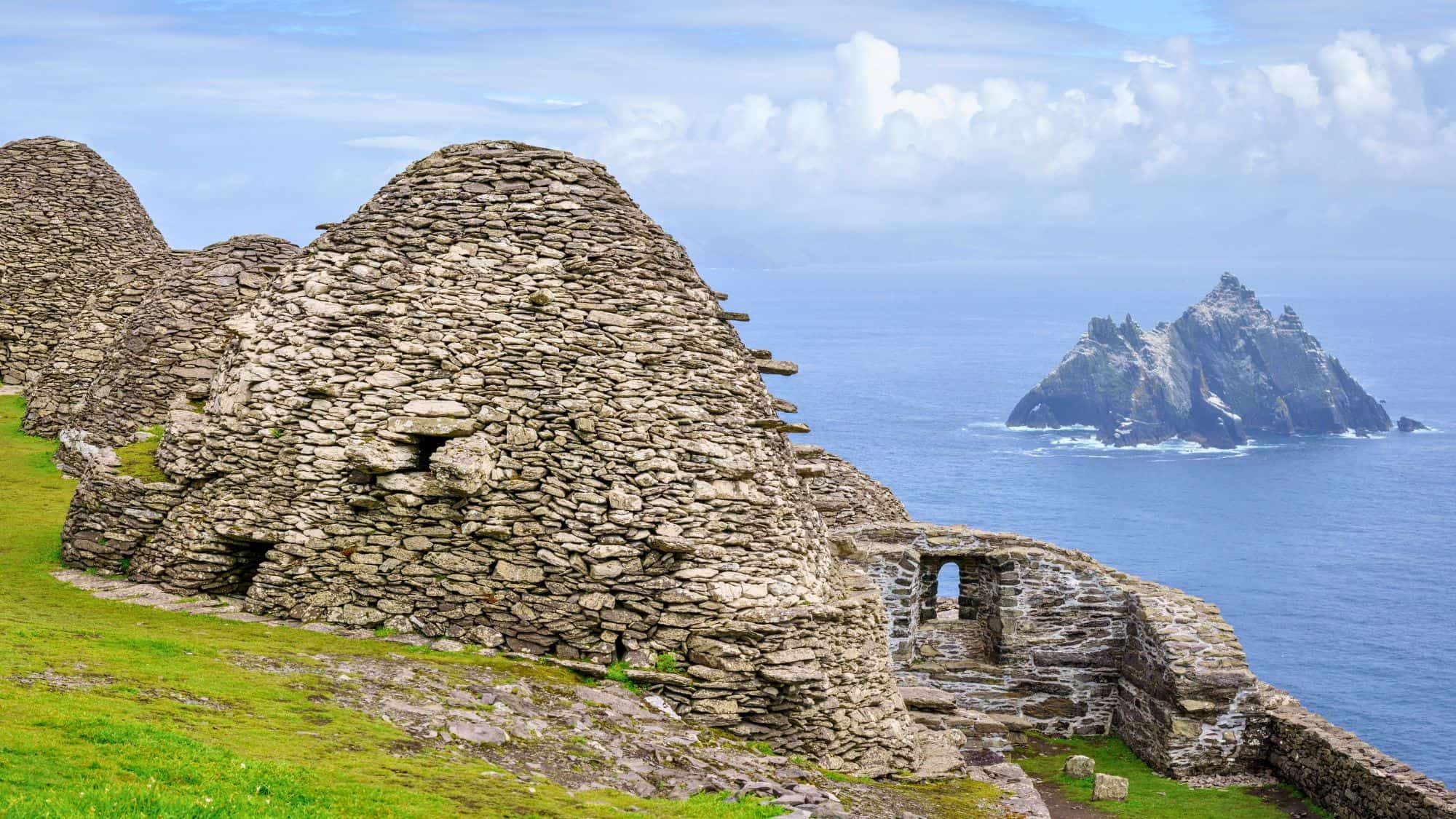 Stone beehive huts perched on a grassy cliffside overlook the dramatic Atlantic Ocean, with Skellig Michael’s iconic jagged rock formation in the distance under a cloudy sky.