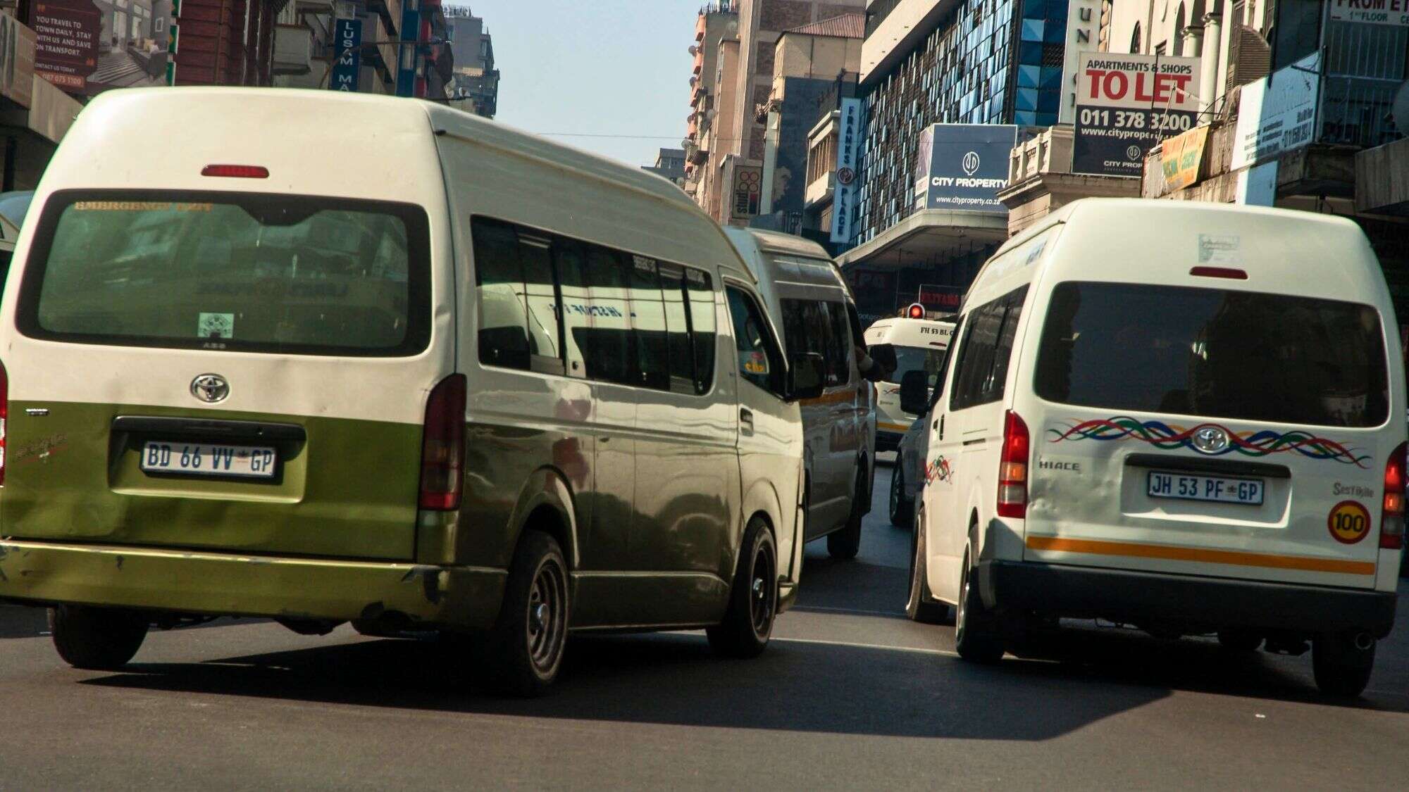 Congested urban street in South Africa filled with white and green minibus taxis, displaying GP (Gauteng Province) license plates and colorful decals.