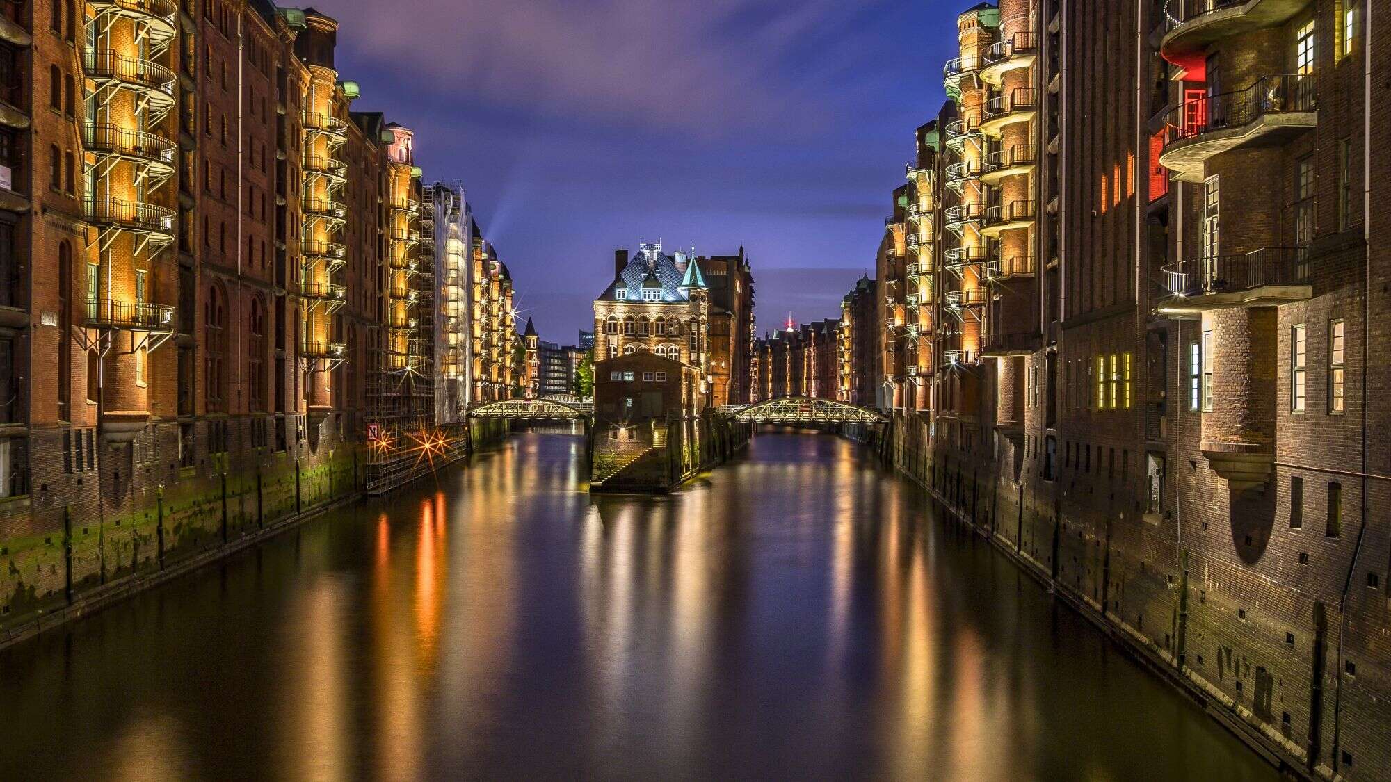 Nighttime image of Hamburg’s Speicherstadt district, with historic brick warehouses illuminated along a canal and a lit-up castle-like building at the canal’s center.