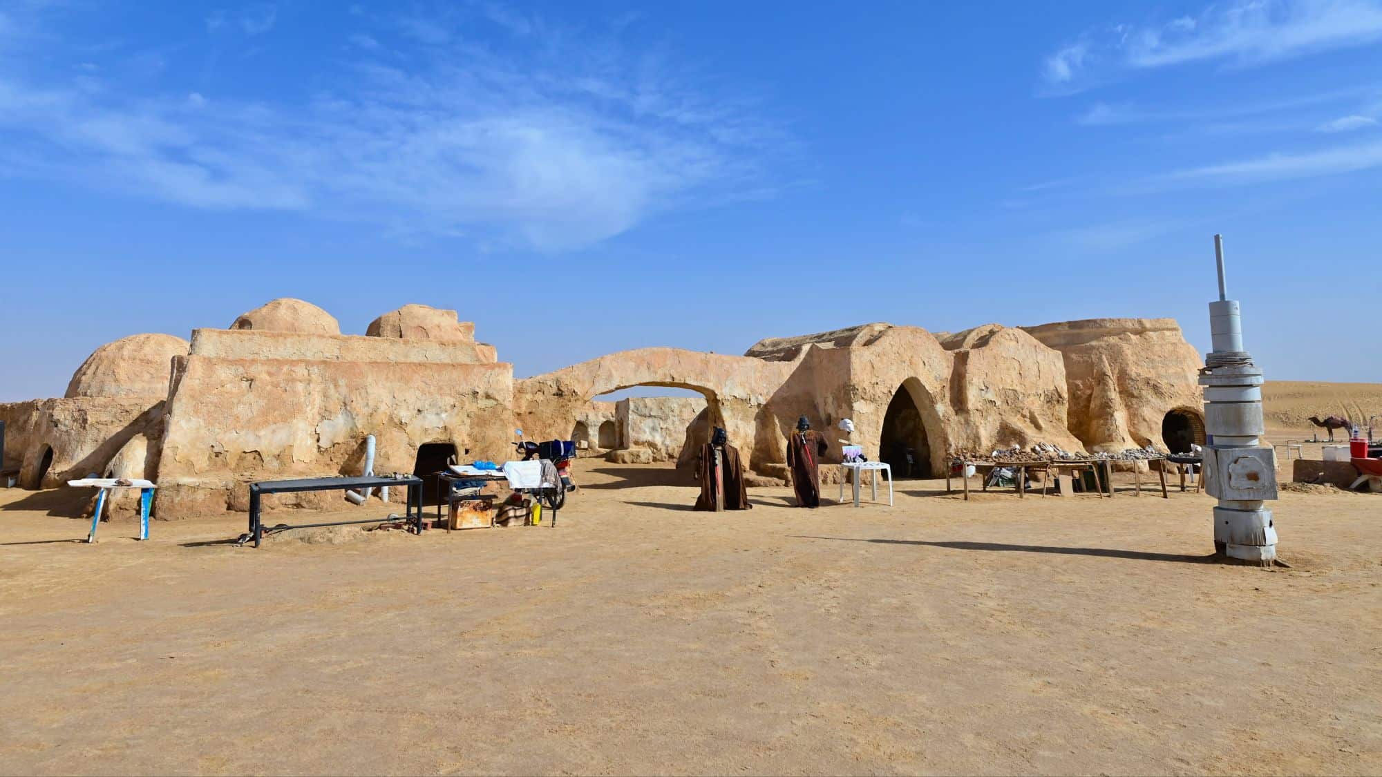 An arid desert scene with domed, sandy-colored buildings and sci-fi props from the Star Wars Tatooine set, surrounded by tables and mannequins dressed as characters from the film.