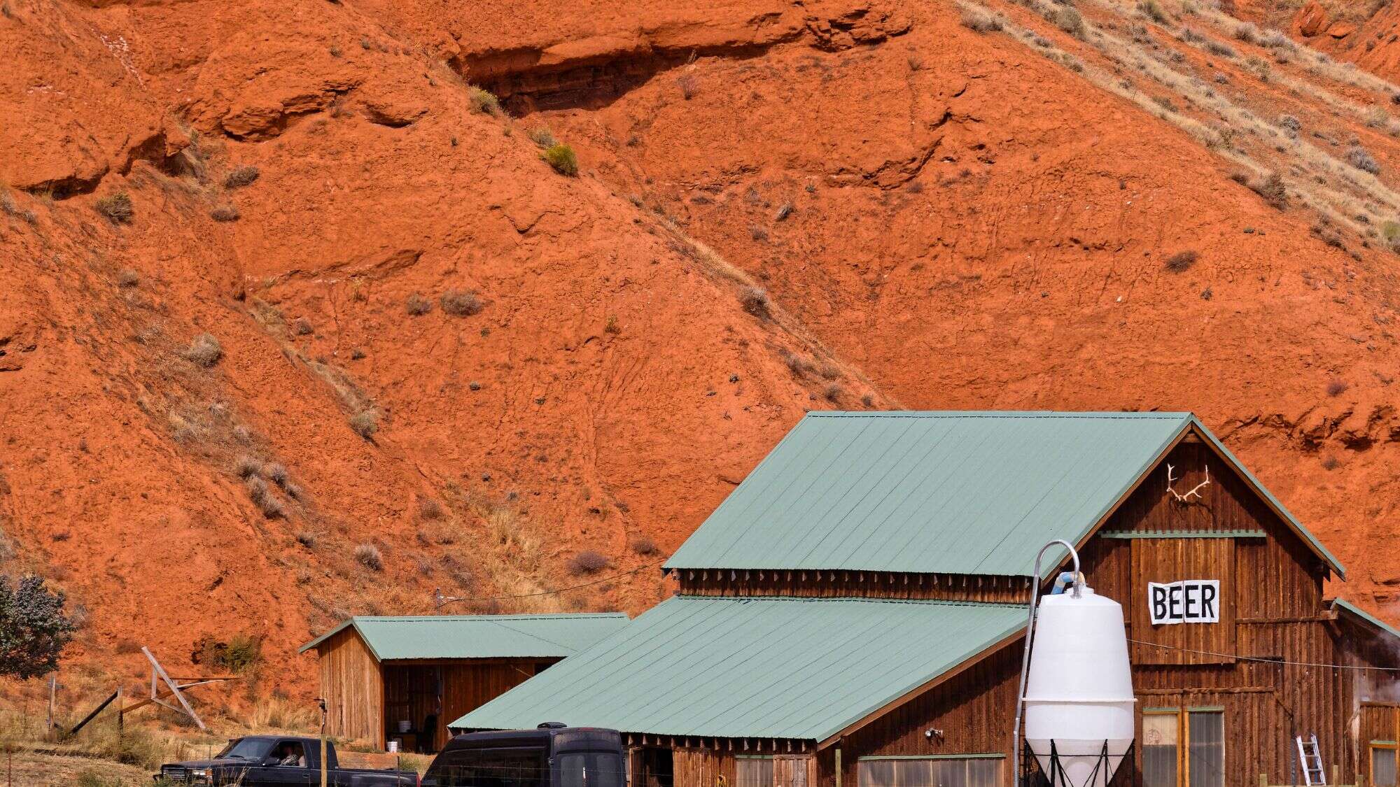 Rustic wooden brewery buildings with green metal roofs stand against vivid red rock cliffs in Ten Sleep, Wyoming, with a sign reading “BEER” and antlers mounted above the entrance.
