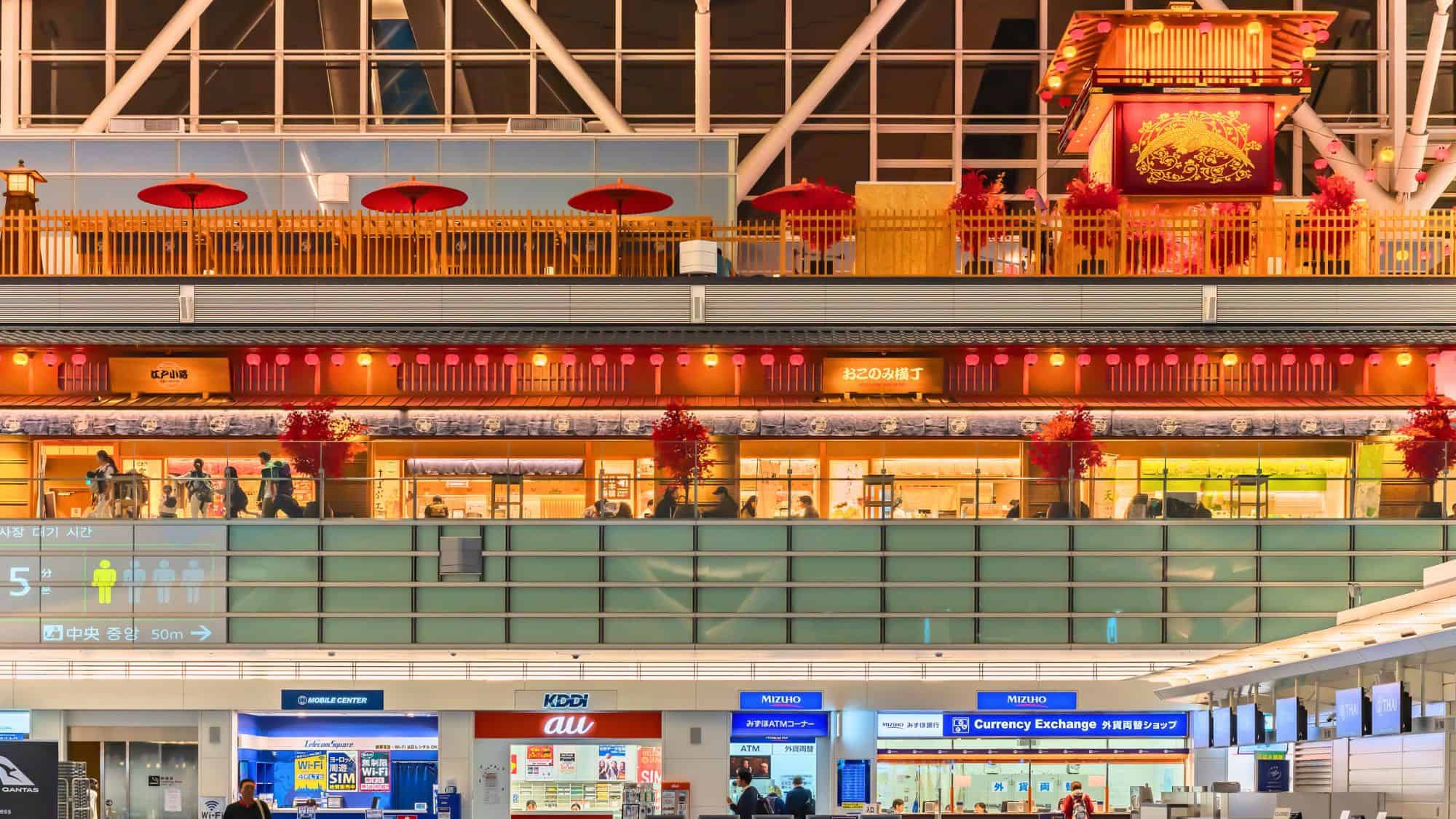 An upper level of Tokyo Haneda Airport mimics an Edo-era Japanese village, with glowing lanterns, red umbrellas, wooden decor, and a modern shopping area beneath it marked by bold signage.