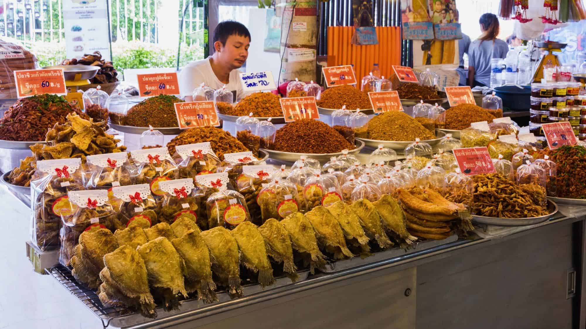 A vendor at Bangkok’s Or Tor Kor Market behind a vibrant spread of dried and spiced fish, chili pastes, and crispy snacks, neatly packaged in bags with red price signs in Thai.