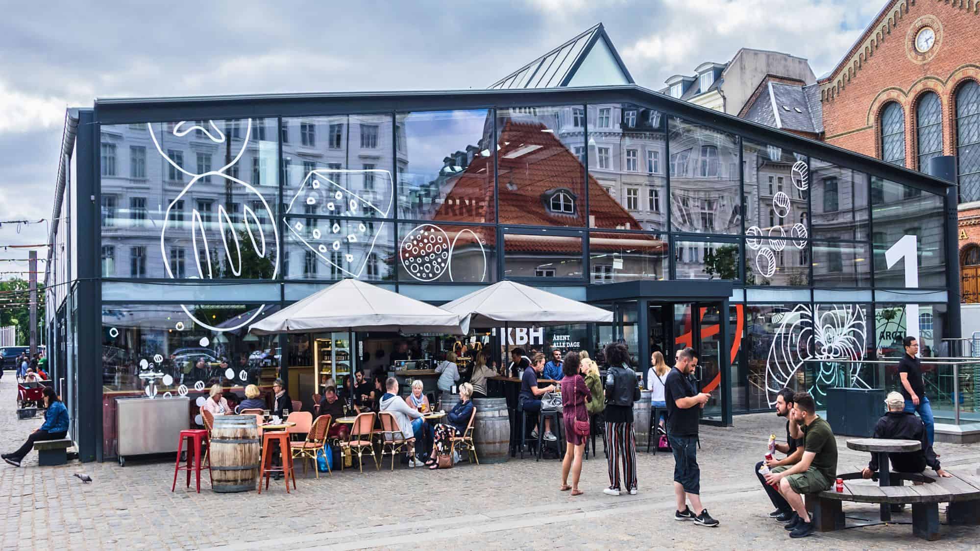 Modern glass market building at Torvehallerne in Copenhagen with outdoor café seating and people enjoying drinks and conversation, surrounded by city architecture reflected in the windows.