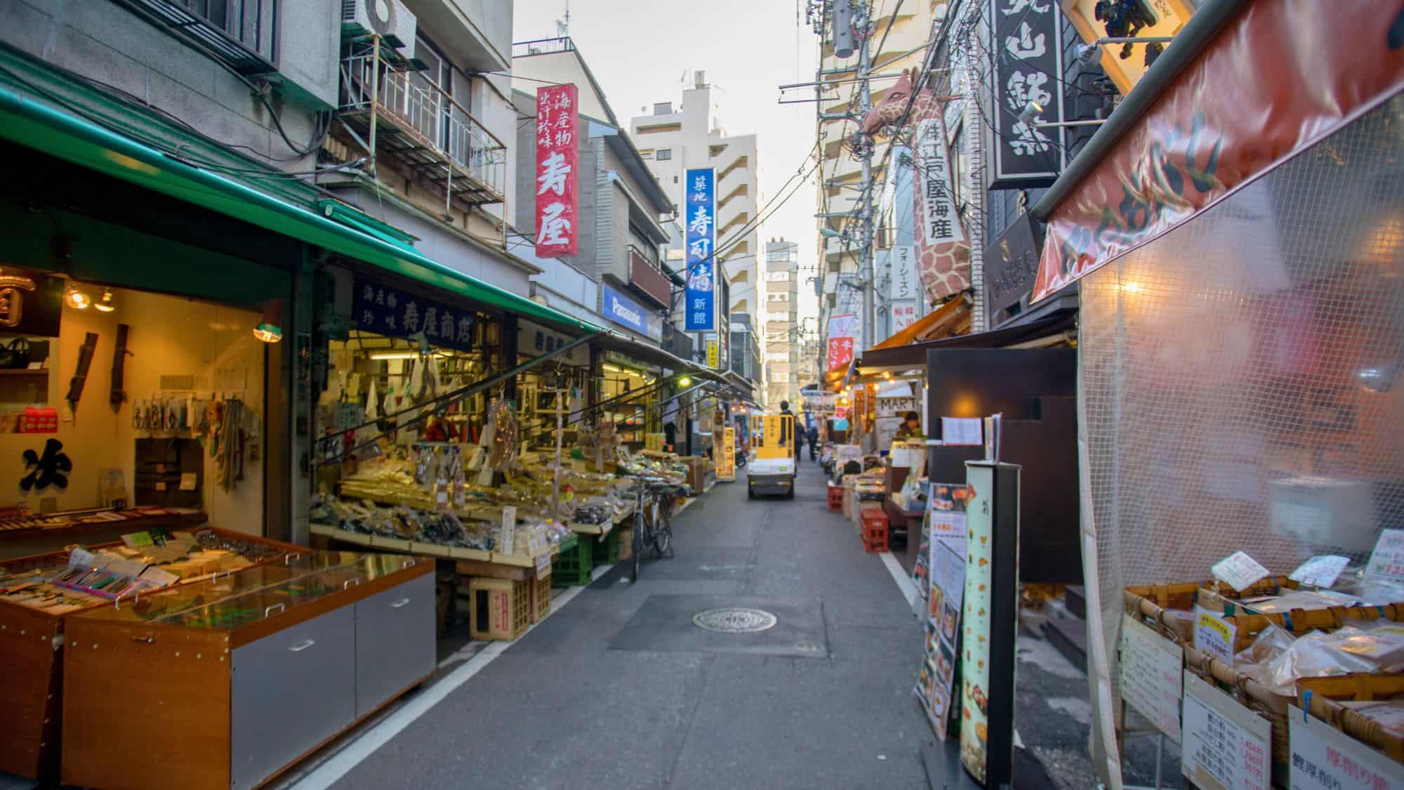Narrow pedestrian street lined with stalls and shops selling seafood, snacks, and kitchen tools at Tsukiji Outer Market in Tokyo, with signs in Japanese and a calm, early morning vibe.