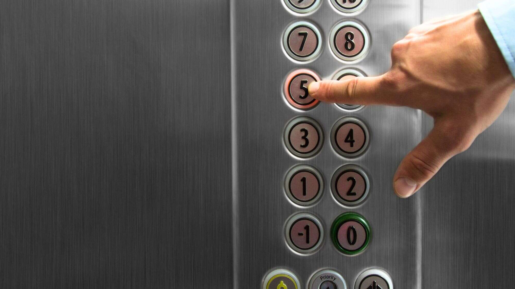 A hand pressing the illuminated button for the 5th floor in a stainless steel UK elevator panel, with buttons numbered from -1 to 8.