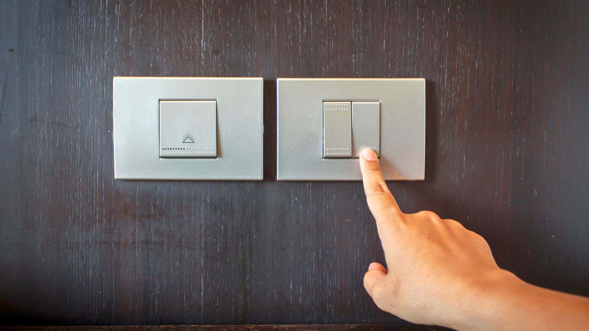 A hand reaching to press a modern square-shaped UK light switch mounted on a dark wooden wall, next to a second identical switch.