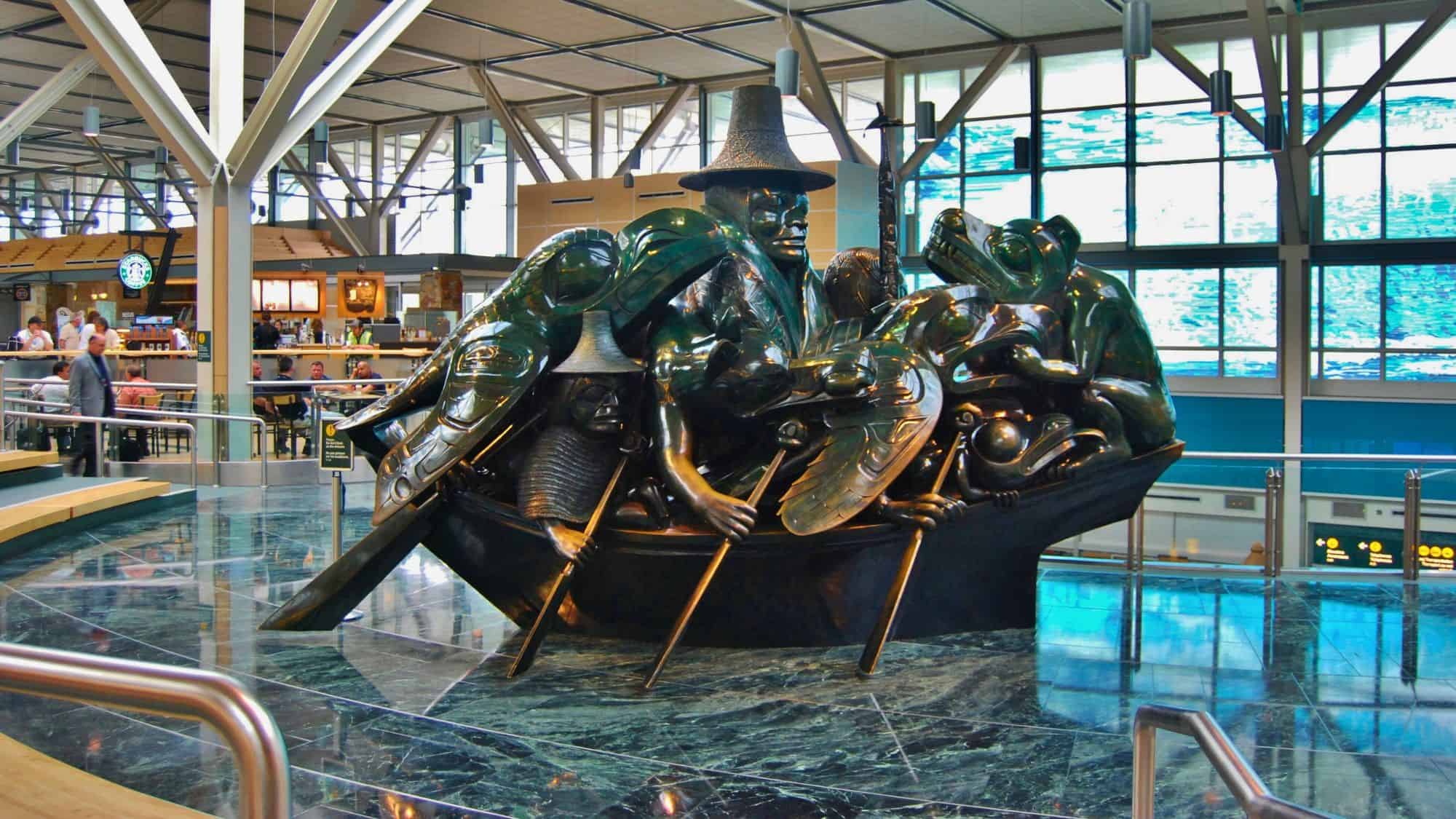 A bronze sculpture titled The Spirit of Haida Gwaii by artist Bill Reid stands at the Vancouver International Airport, featuring a canoe packed with figures from Haida mythology under bright terminal lighting and surrounded by travelers.